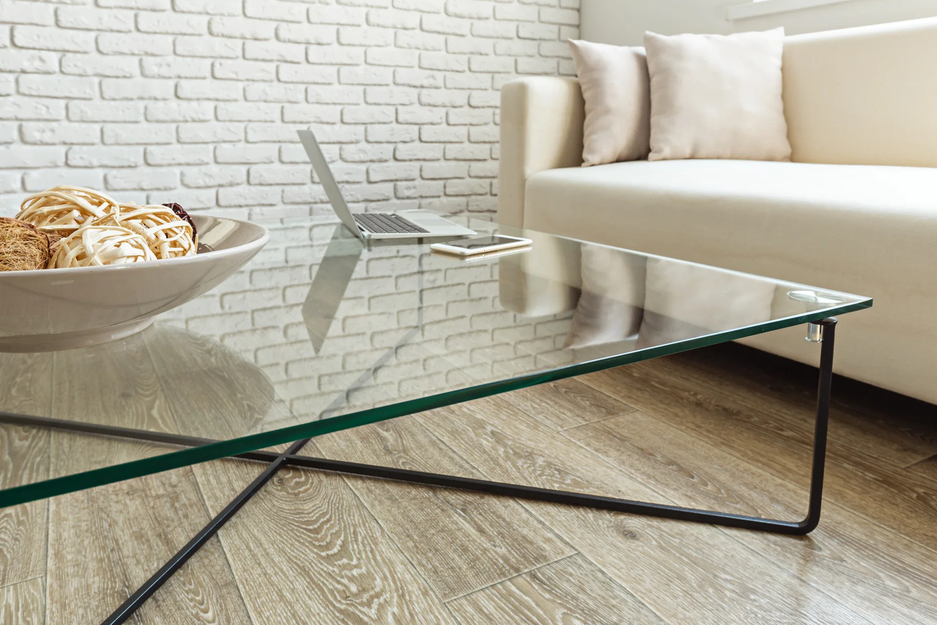 A glass-topped coffee table with a black metal frame sits in front of a cream sofa and a white brick wall.