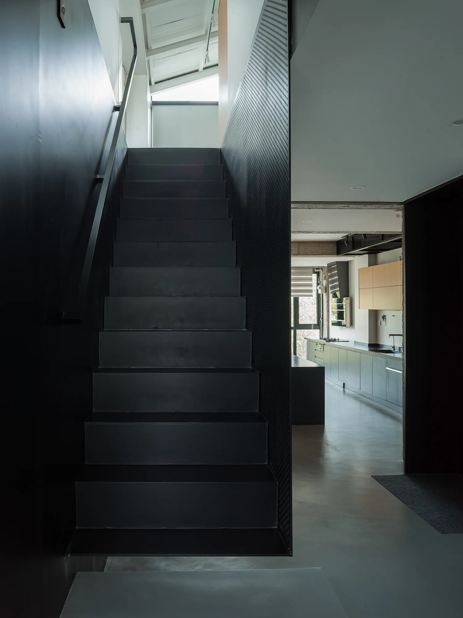A matte black metal staircase with a perforated screen panel rises from a smooth gray floor next to a modern kitchen in the background.