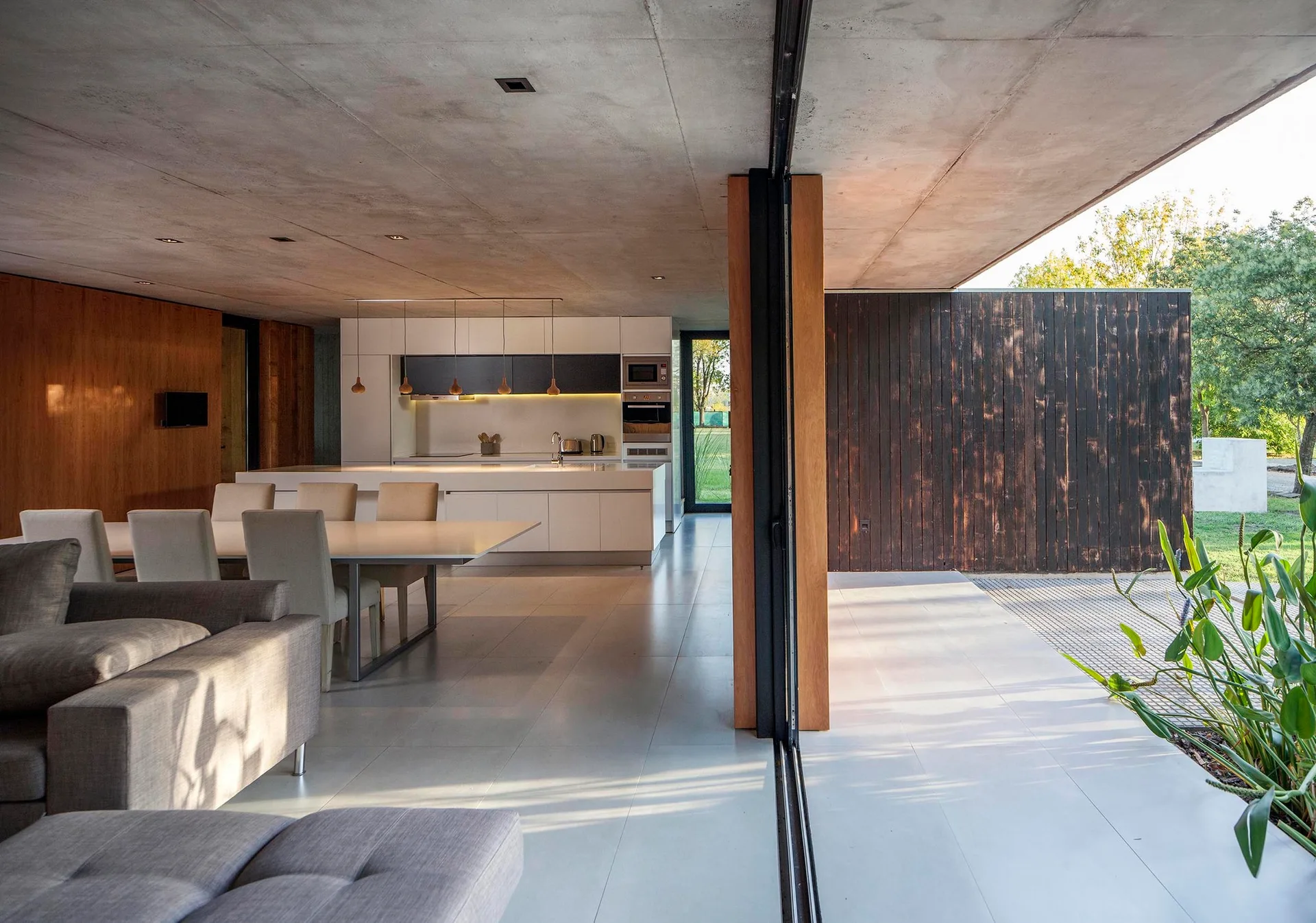 An open-plan living room and kitchen featuring a grey sofa, dining table, and minimalist white cabinets beneath an exposed concrete ceiling.