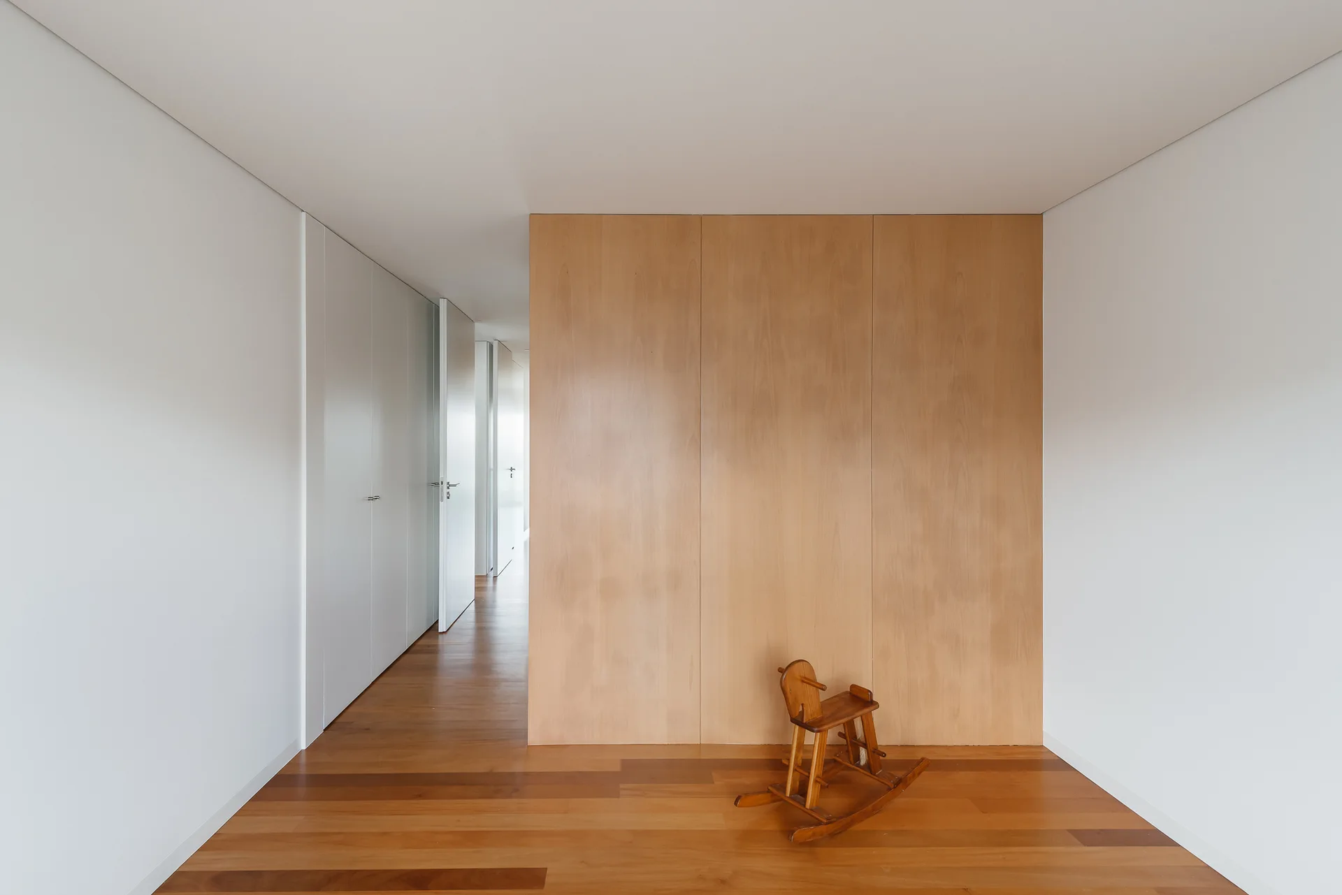 A minimalist hallway featuring white walls, a central light wood partition, wood plank floors, and a small wooden rocking horse.