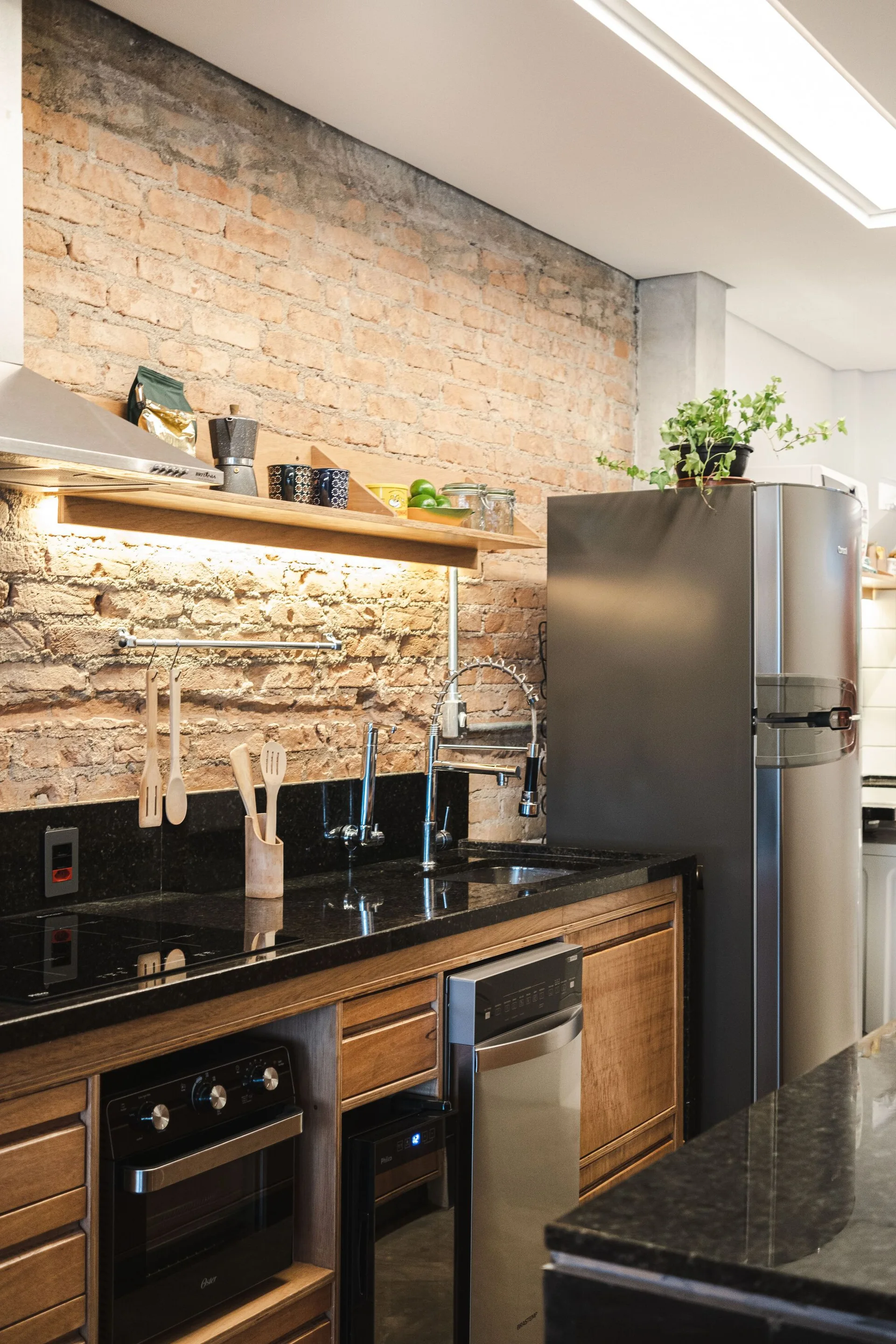 A kitchen featuring an exposed brick wall, a black countertop with an undermount sink, wood base cabinets, and a stainless steel refrigerator.