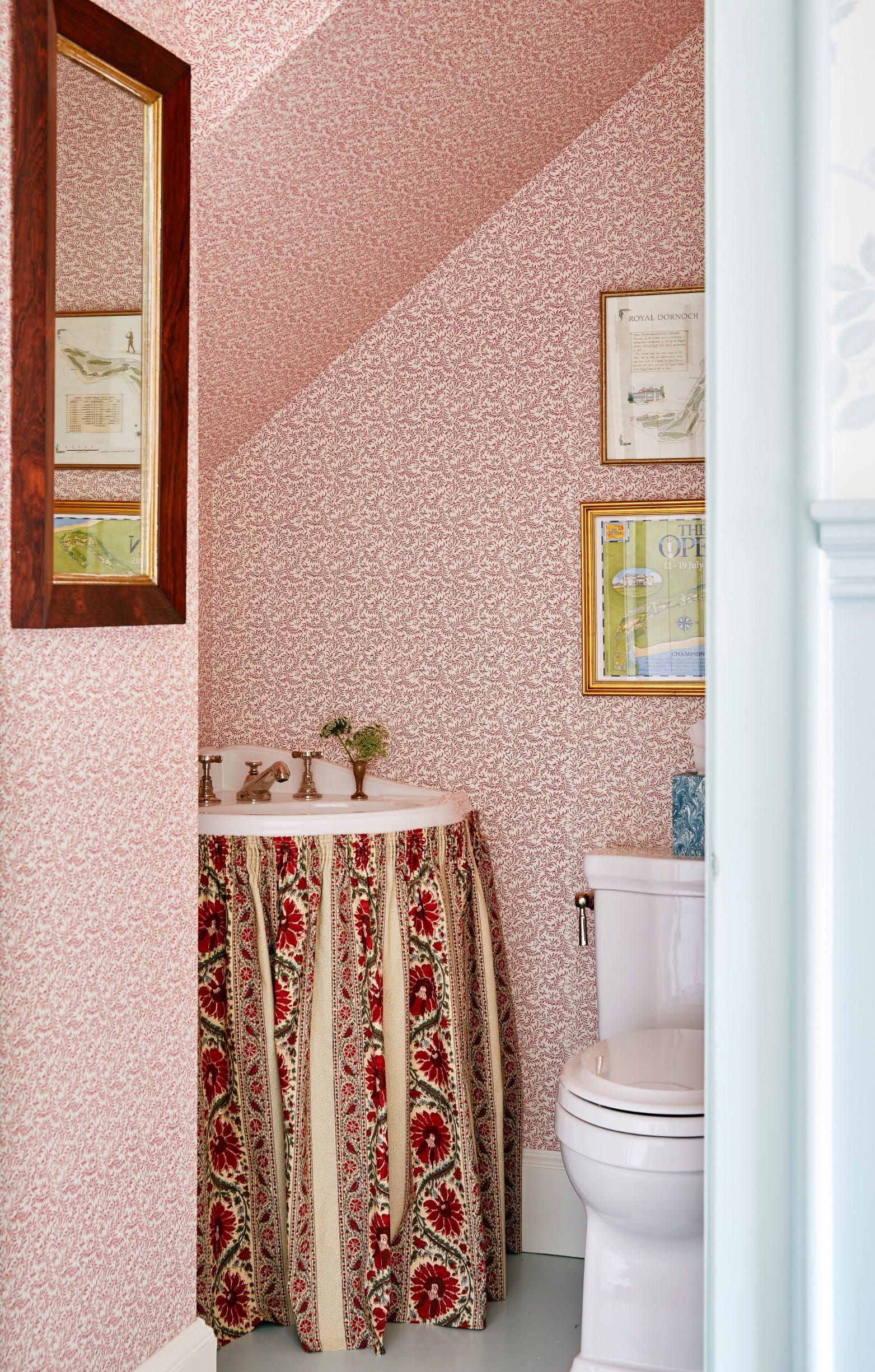 A small powder room features red botanical wallpaper, a white toilet, and a sink concealed by a gathered floral fabric skirt.