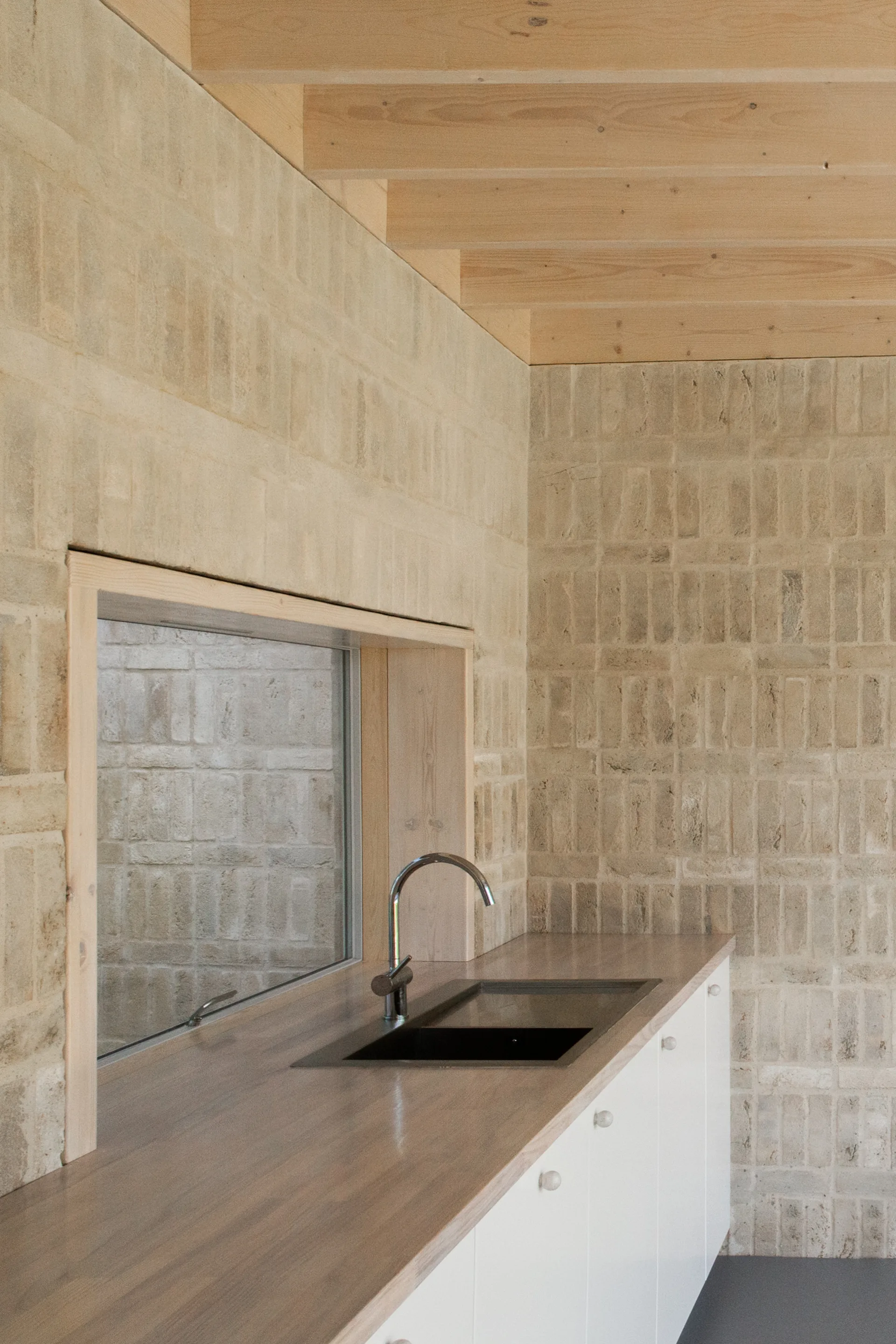 A modern kitchen featuring beige brick walls, exposed wood ceiling beams, a wood countertop with an inset black sink, and white lower cabinets.
