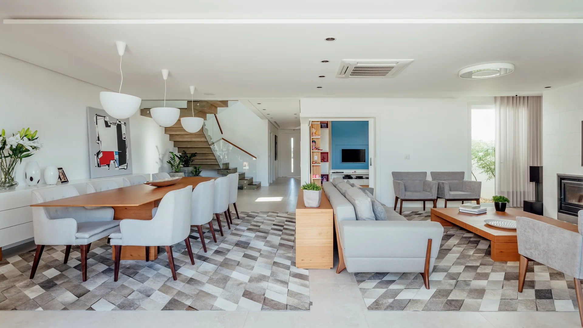 An open-concept living and dining room features a wooden dining table, a grey sofa, matching checkered area rugs, and a wooden floating staircase.