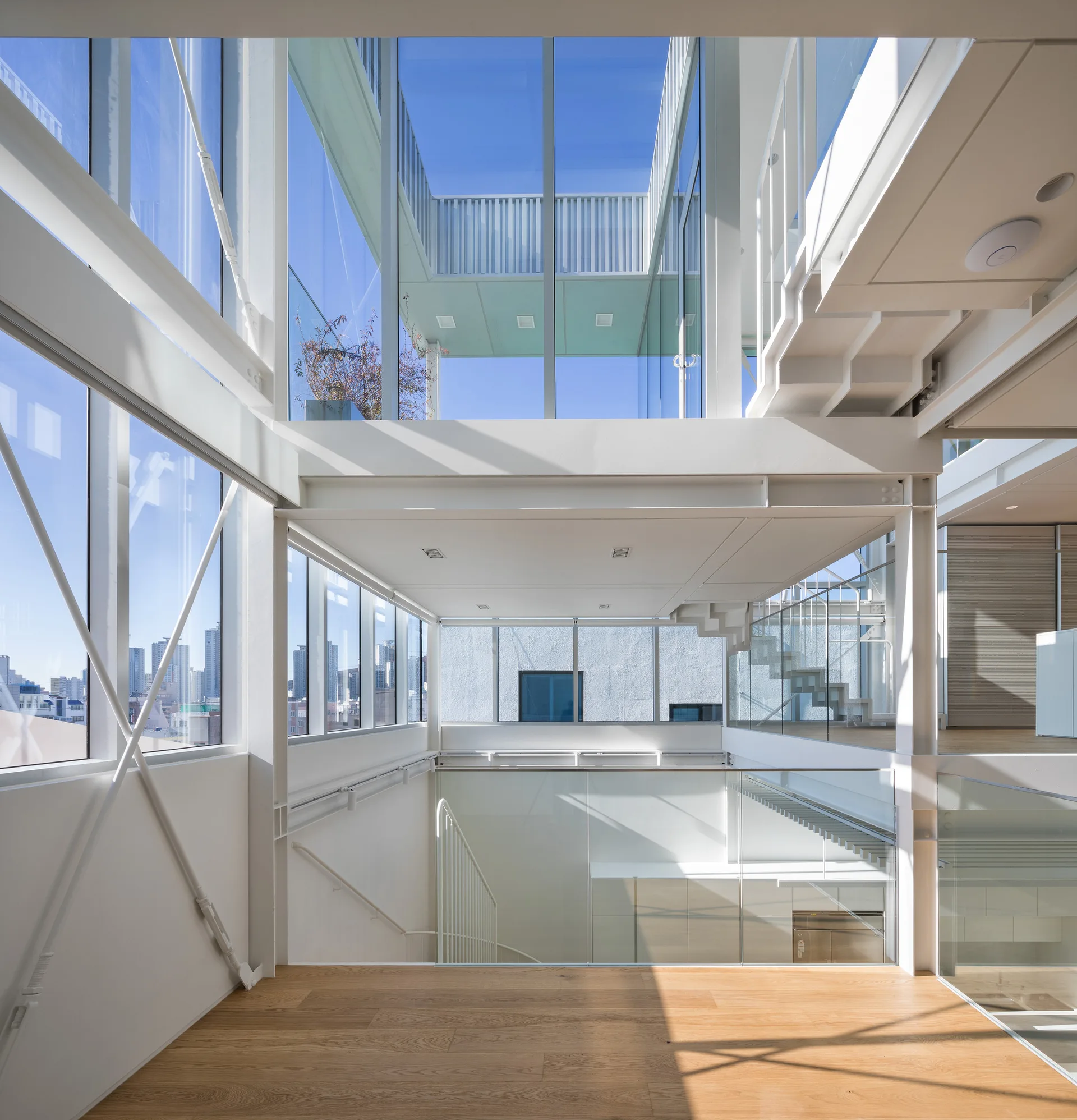 A multi-level modern interior featuring white steel structural beams, floor-to-ceiling glass walls, an open staircase, and light wood flooring.