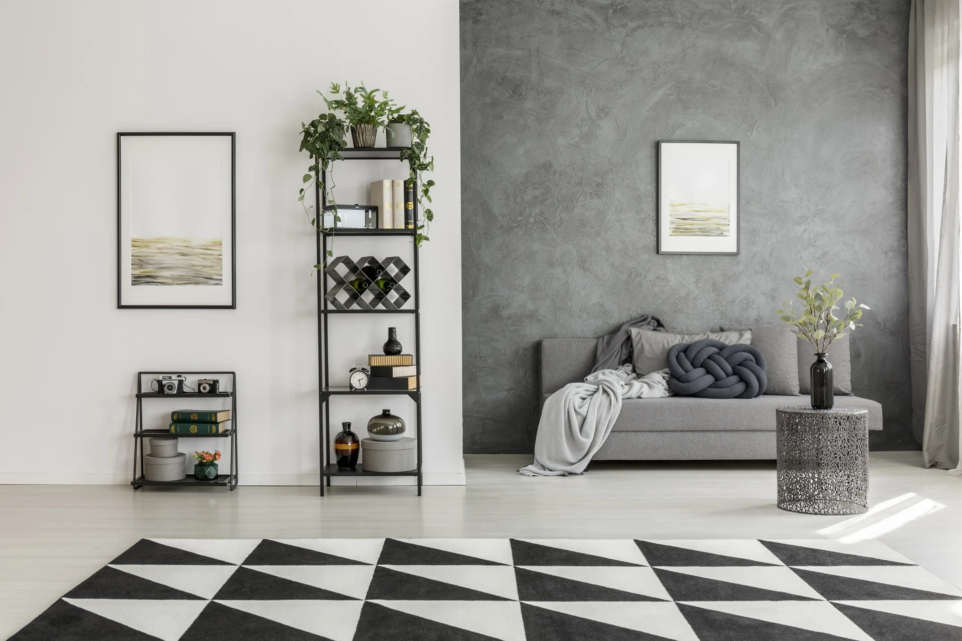 A grey sofa sits against a textured grey accent wall in a living room featuring a geometric black and white rug and a tall black metal shelving unit.