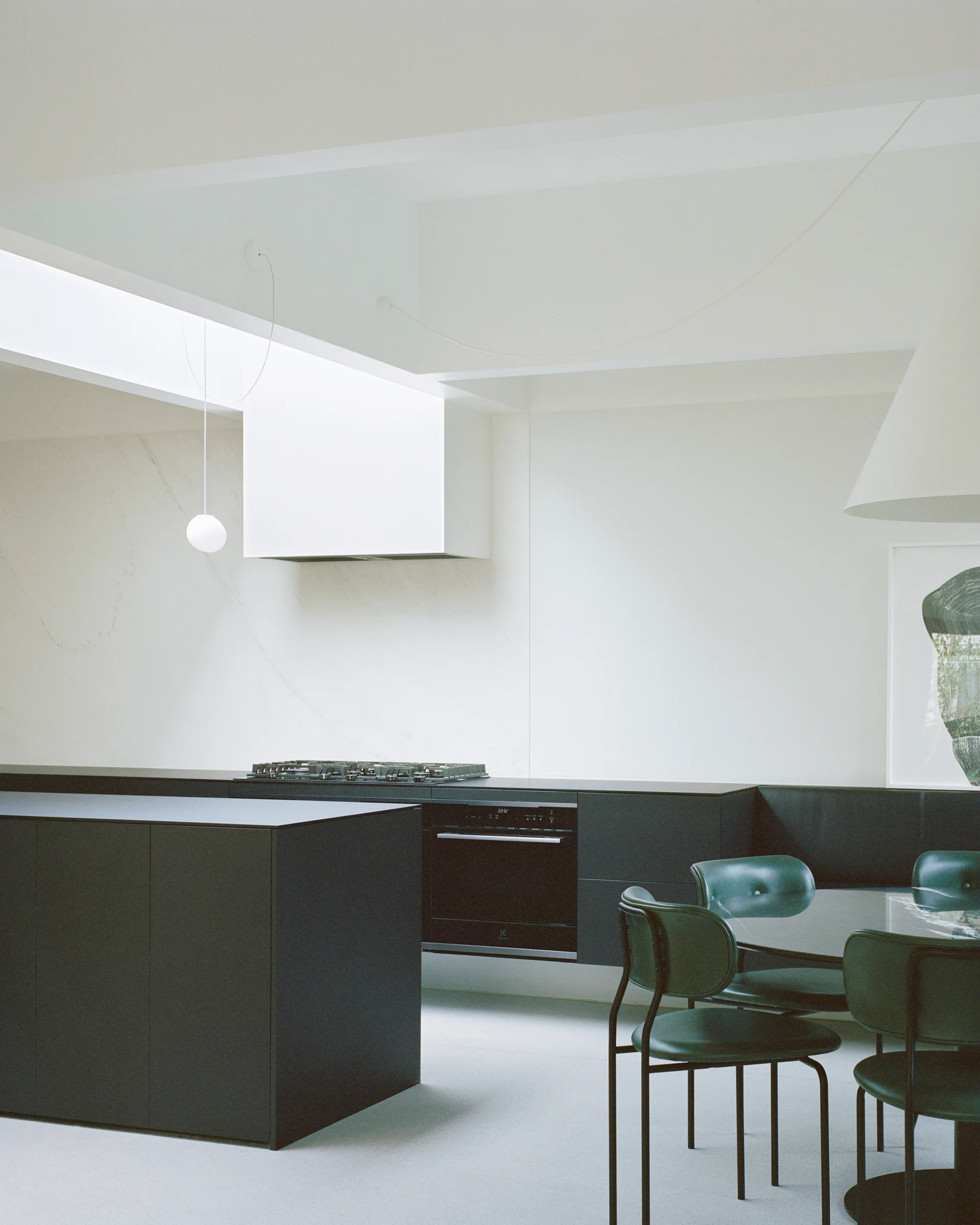 A minimalist kitchen features a dark charcoal island and matching lower cabinetry next to a dining area with a round table and forest green chairs.