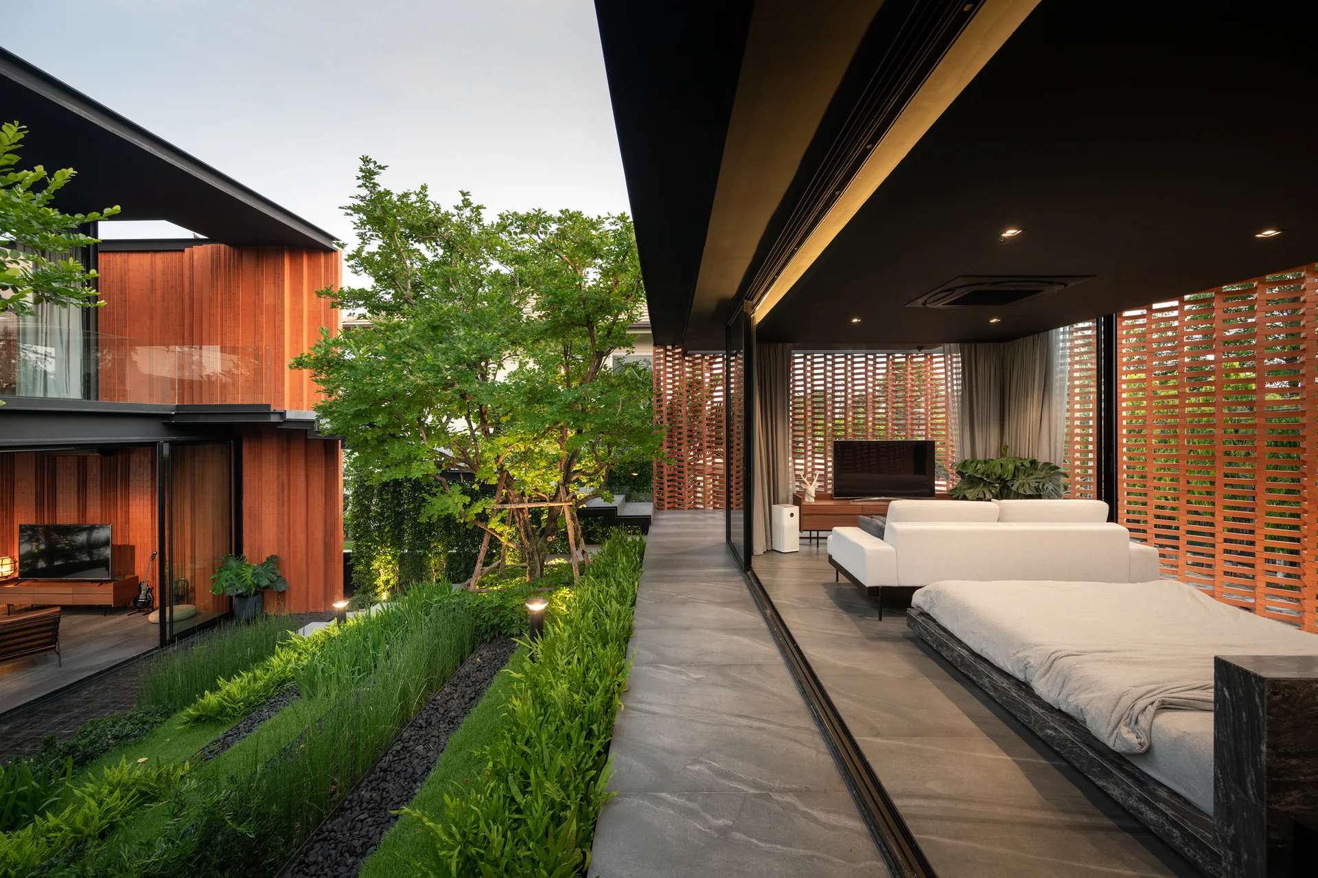A minimalist bedroom with a platform bed and white sofa opens onto a landscaped garden through sliding glass doors under a dark ceiling.
