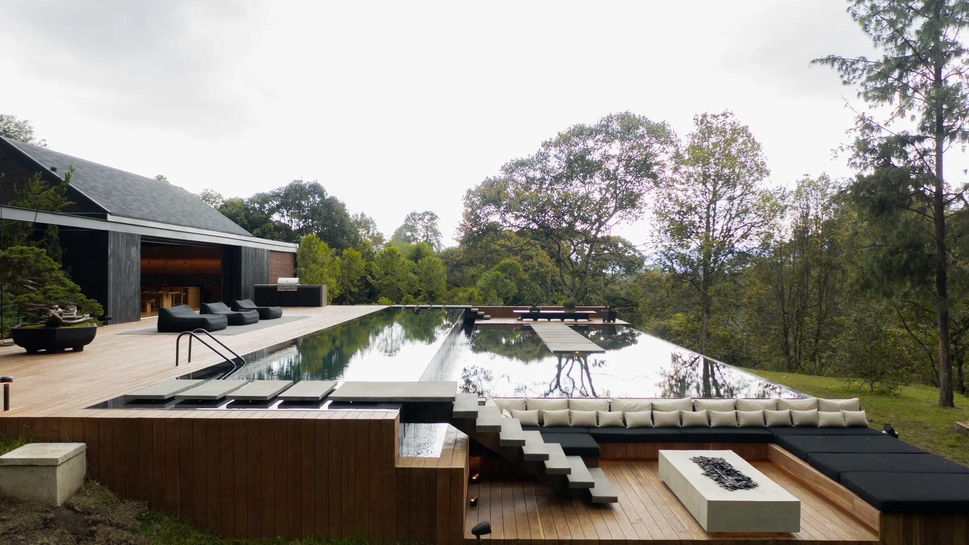 A modern outdoor wood deck features an infinity pool, a sunken seating area with black cushions, a concrete fire pit, and floating stone steps.