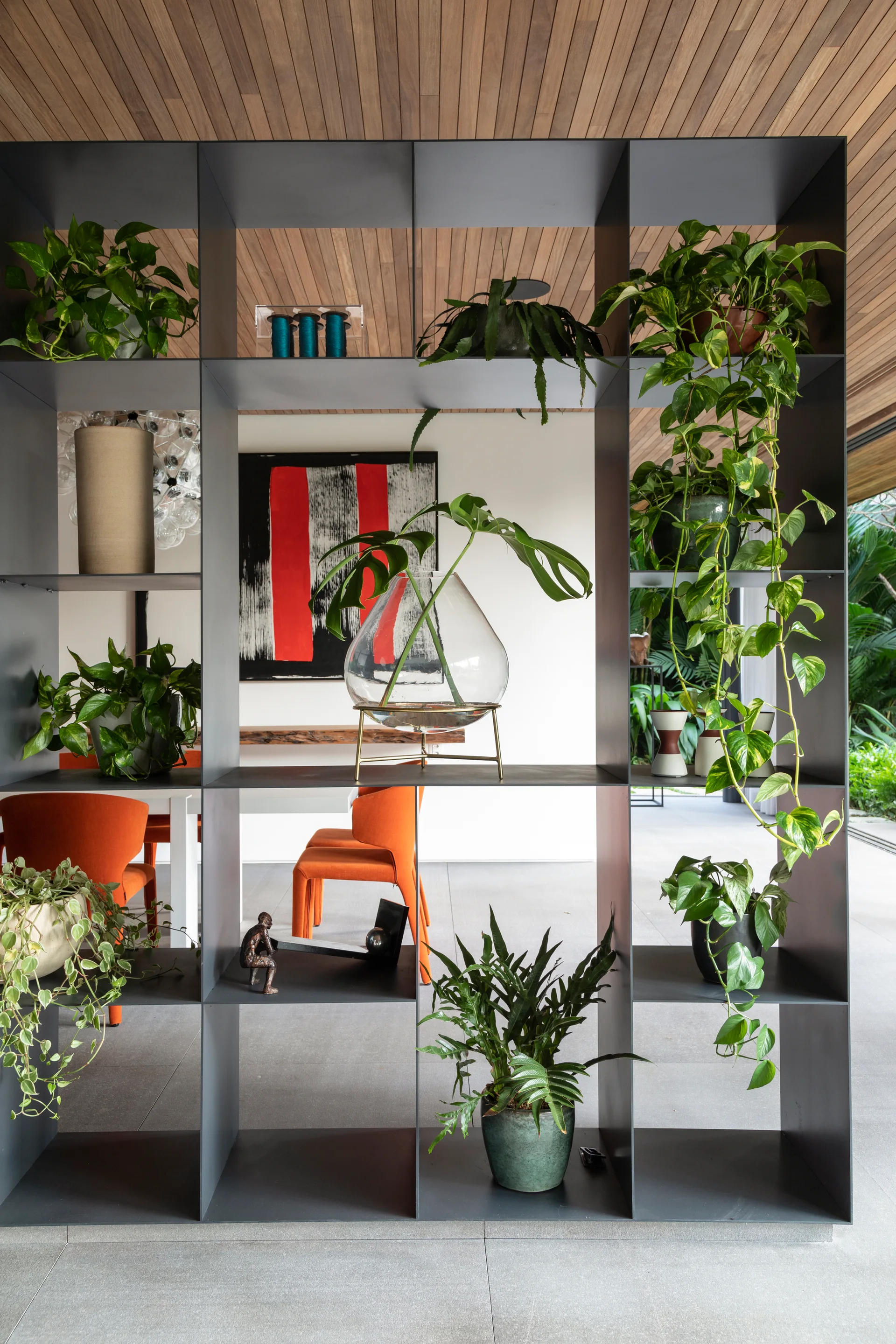 A dark grey metal open shelving unit filled with potted plants divides a modern room featuring a wood slatted ceiling and orange dining chairs.