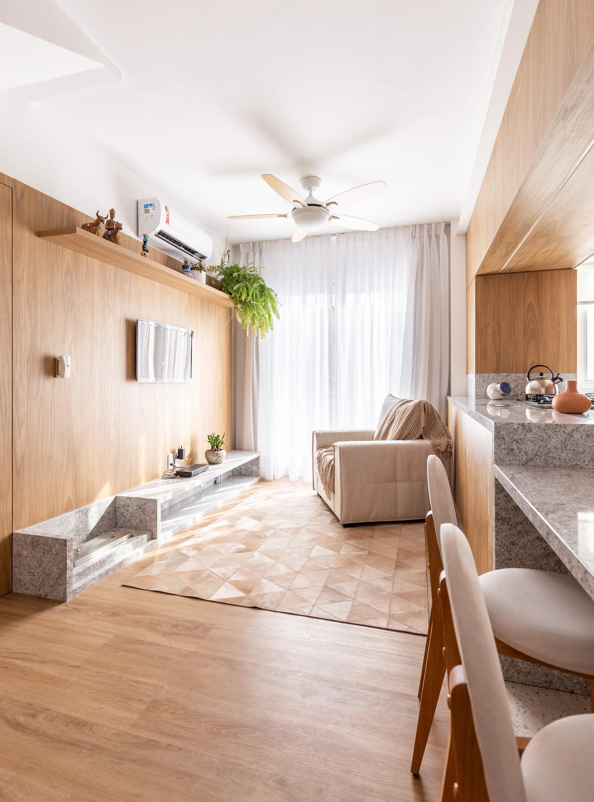A compact living room features a wood-paneled TV wall with a low stone-like console, a geometric patterned rug, and an adjacent kitchen bar.