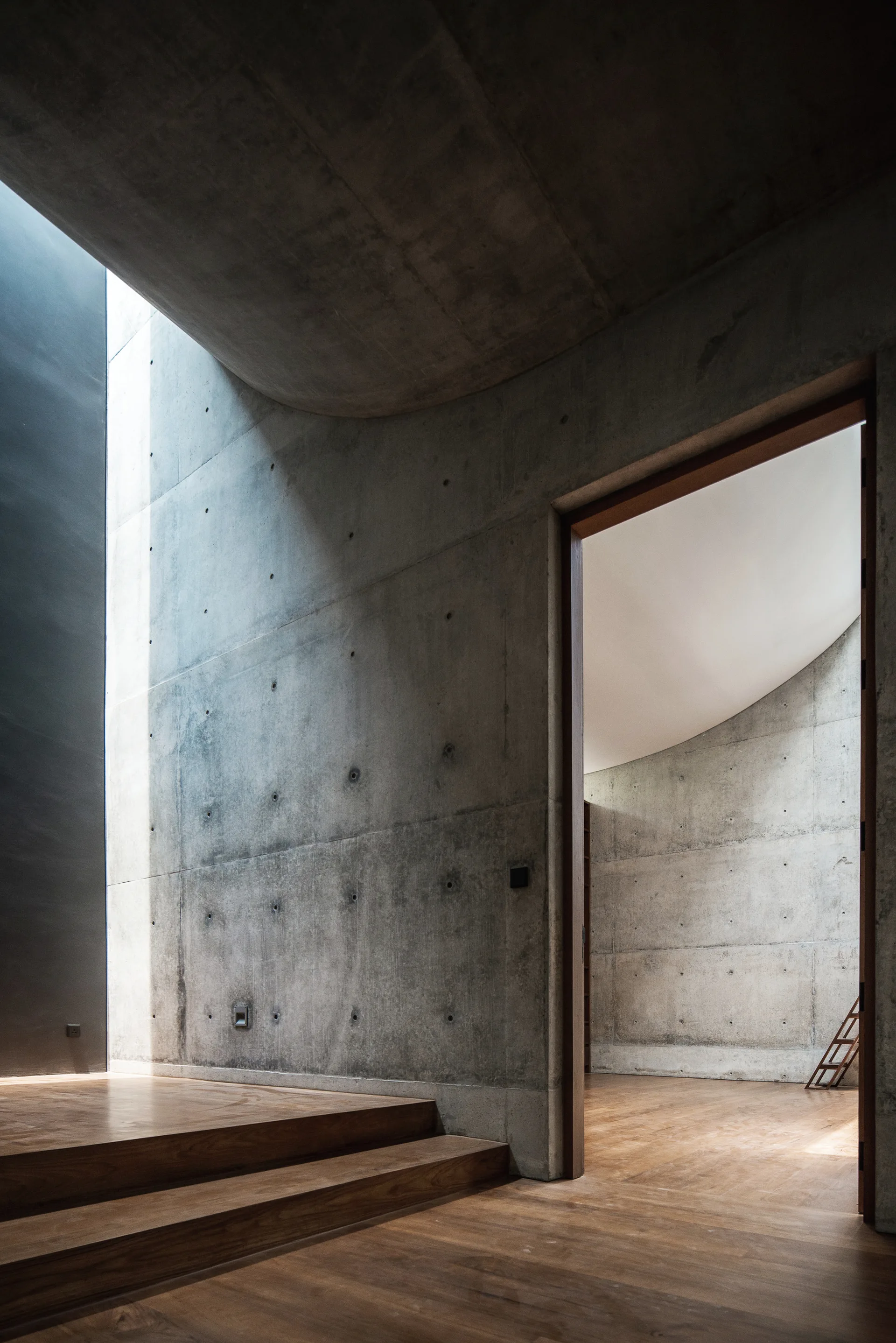 A minimalist interior corridor features raw concrete walls with tie rod holes, low wooden steps, and a doorway leading to an adjacent room.