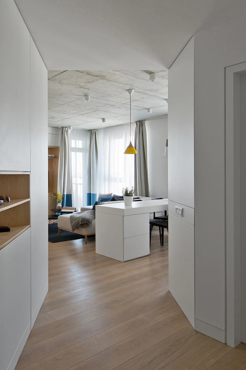 A modern apartment hallway looking toward a white dining table, an exposed concrete ceiling, and a light wood floor.