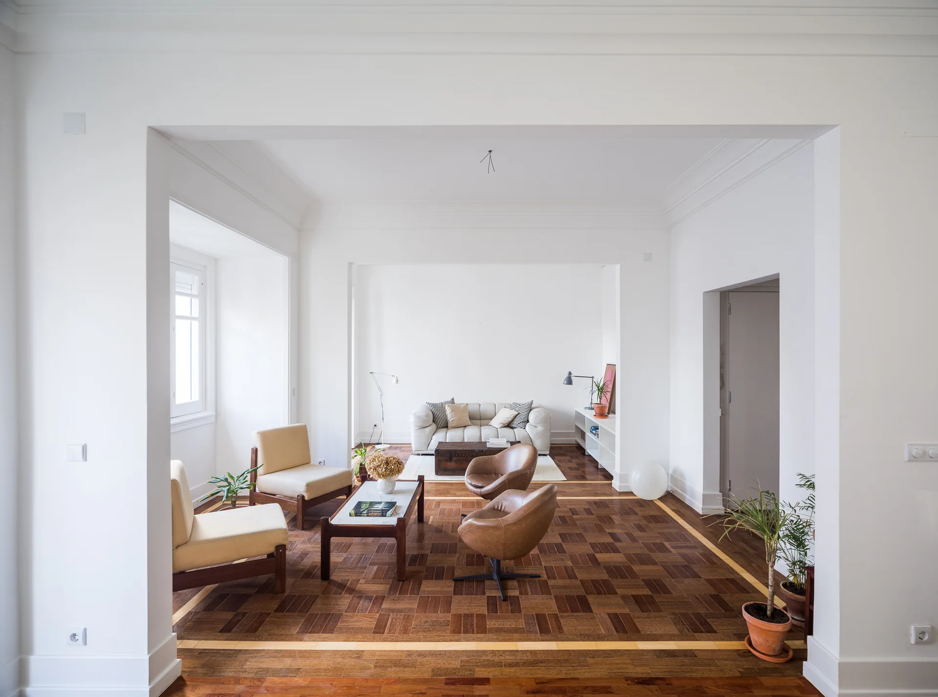 A spacious white living room with patterned parquet wood flooring features beige armchairs, brown leather swivel chairs, and a white sofa in the background.