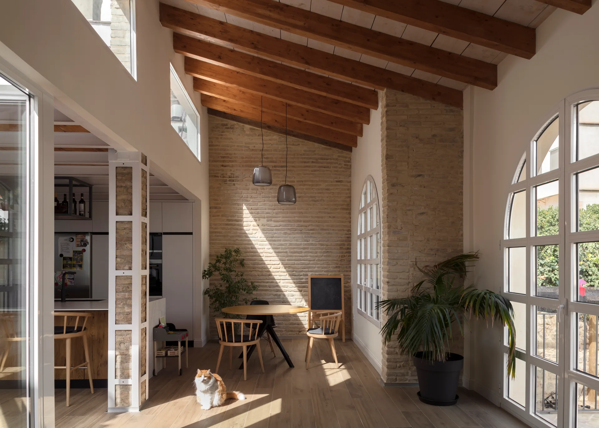 A dining room features a round wooden table, spindle chairs, exposed brick walls, and a sloped ceiling with rustic wood beams.