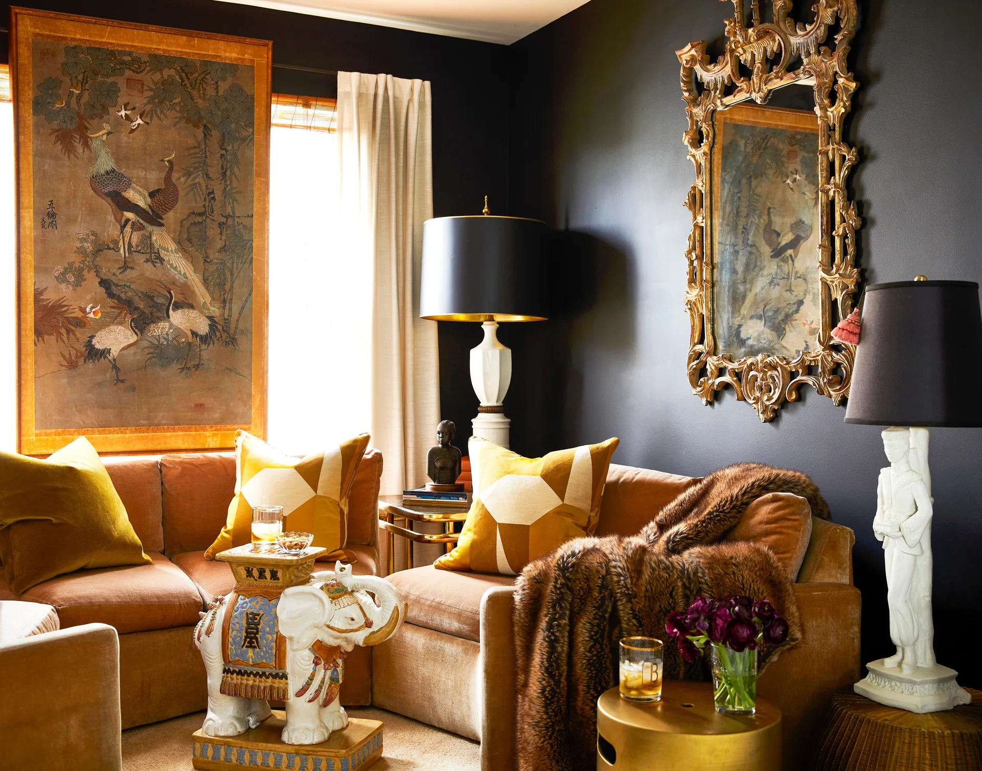 A tan velvet sectional sits against dark walls, styled with an ornate gold mirror, white figural lamps, and a ceramic elephant side table.