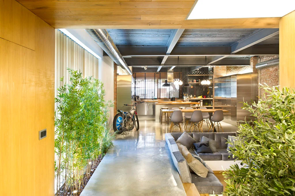 An industrial loft interior featuring a polished concrete floor, a tall indoor bamboo planter, a grey sectional sofa, and exposed steel ceiling beams.