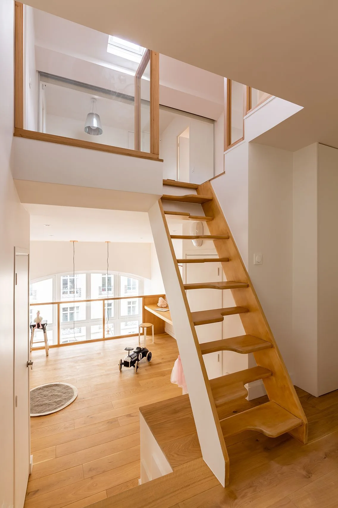 A wooden staircase leads to a mezzanine level in a white loft apartment with a large arched window and light wood floors.