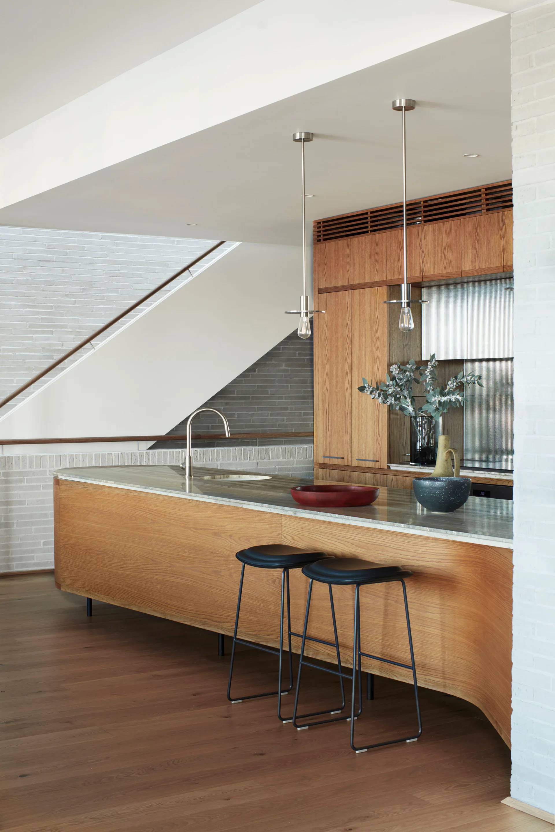 A modern kitchen featuring a curved wood island with a stone countertop, black bar stools, and tall integrated wood cabinetry.