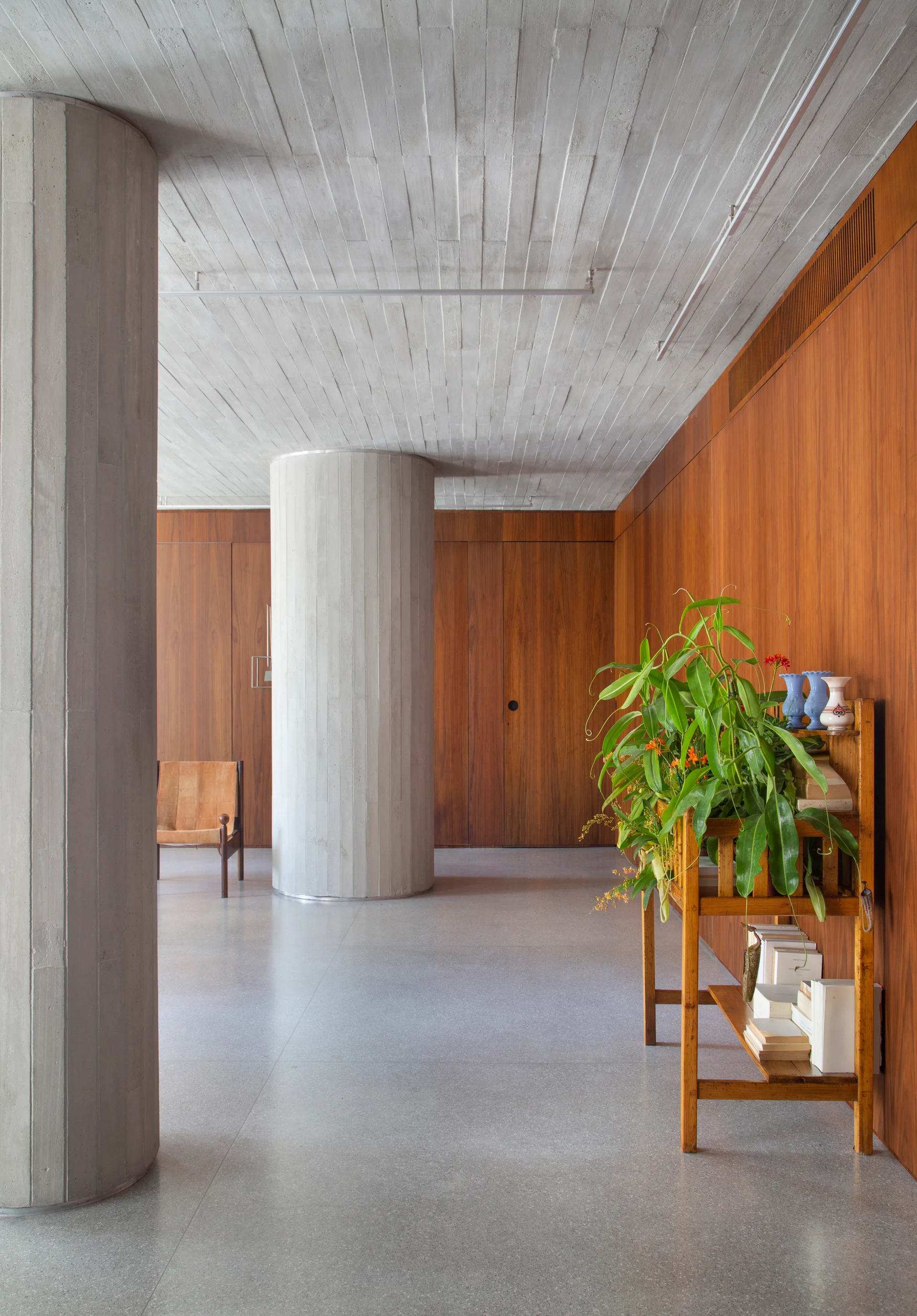 A spacious minimalist interior featuring prominent board-formed concrete columns, a matching concrete ceiling, and warm vertical wood wall paneling.