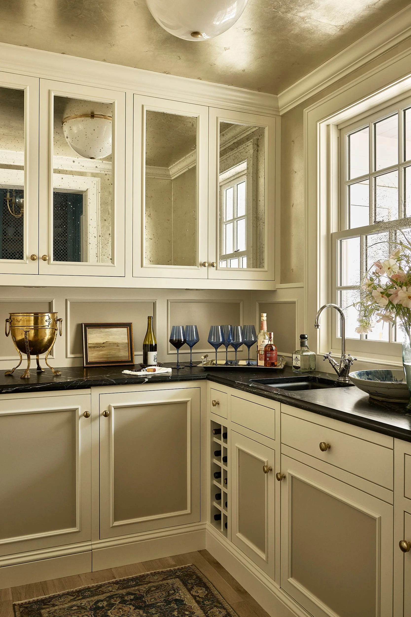 Cream butler's pantry features antique mirrored cabinet doors, dark veined stone countertops, a small sink, and a metallic gold leaf ceiling.