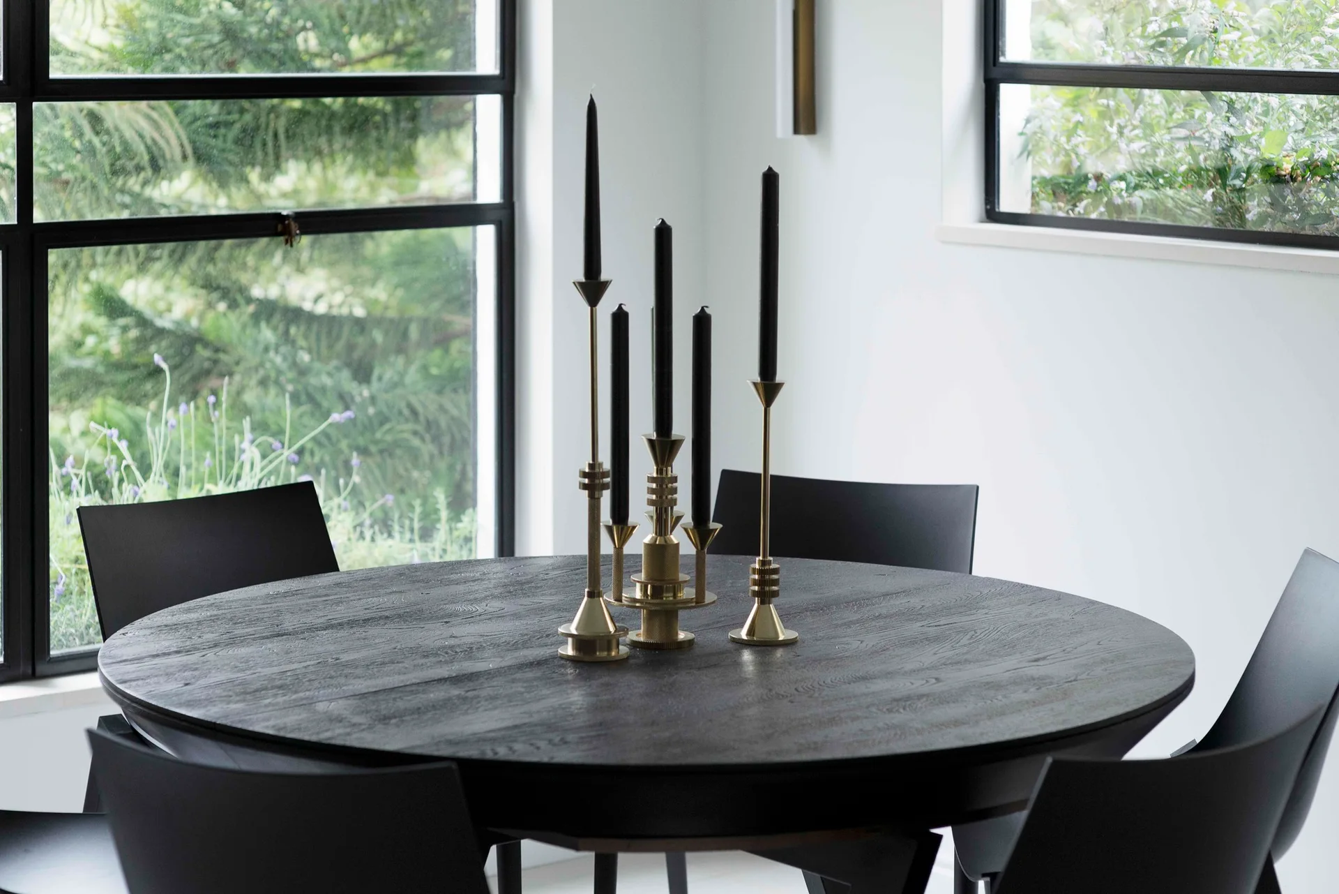 A round black wood dining table with minimalist chairs features a gold brass candelabra centerpiece holding black taper candles.
