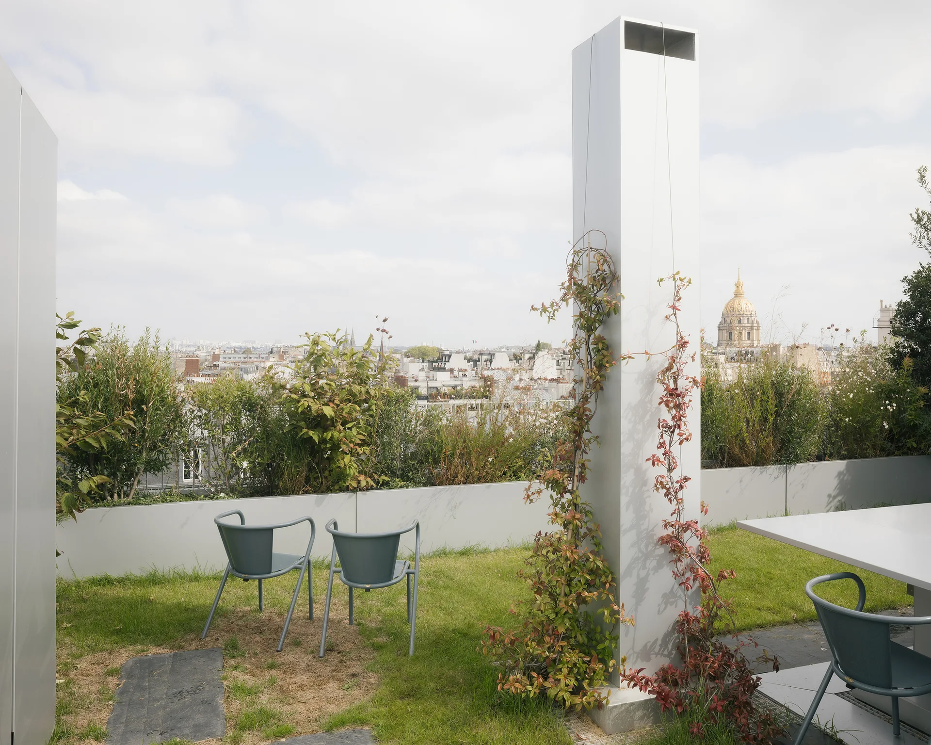 A minimalist rooftop terrace features a green grass lawn, gray metal chairs, planter boxes with lush shrubs, and a tall metal column with climbing vines.