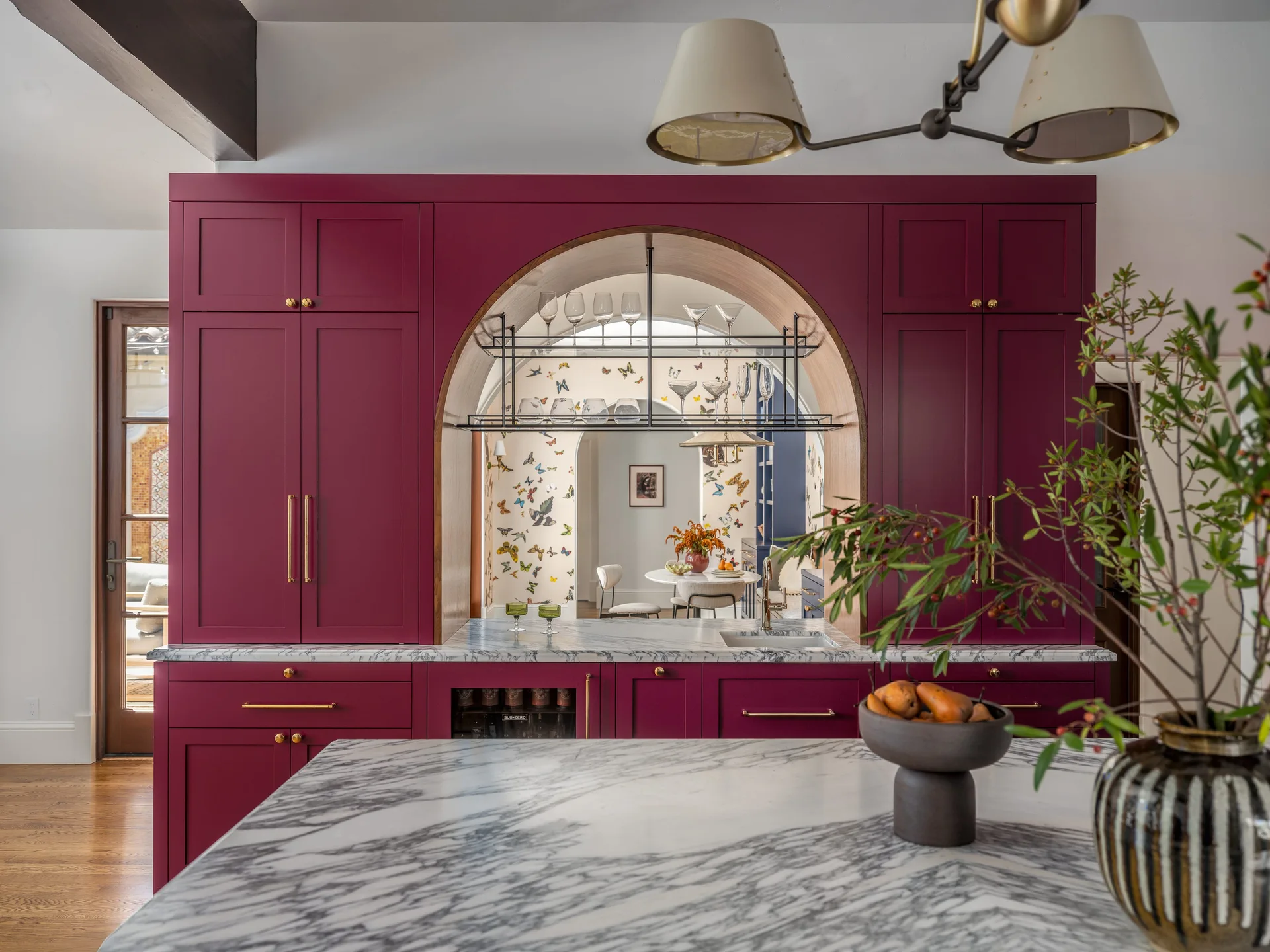 Deep magenta kitchen cabinetry features an arched cutout with a black metal hanging rack, showing butterfly wallpaper in the room behind.