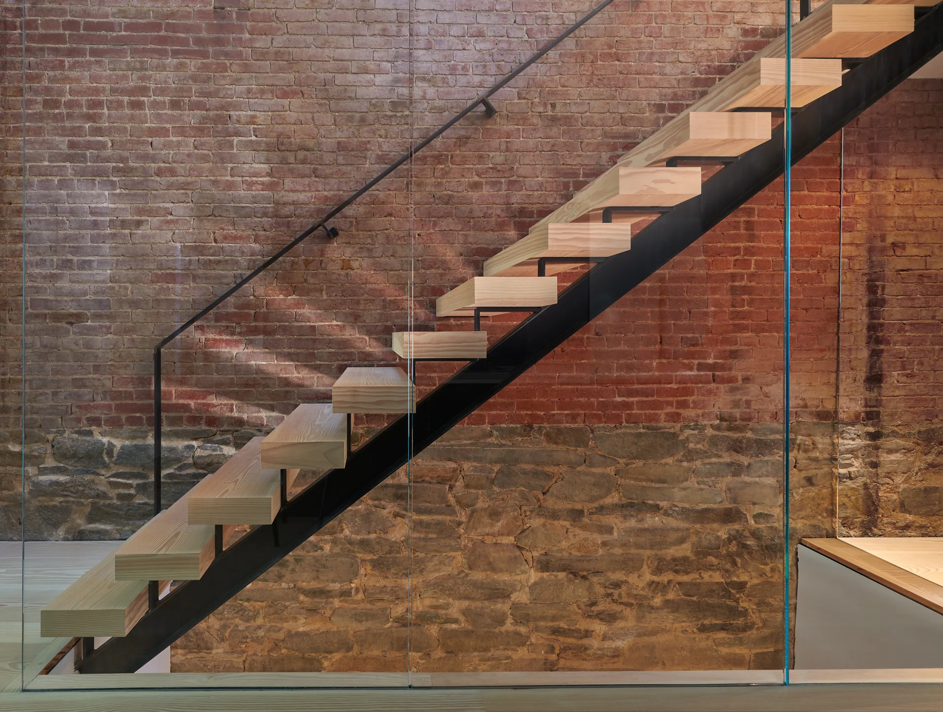 A modern staircase with thick wood treads and a black metal frame sits next to a glass partition against a background of red brick and rough stone.
