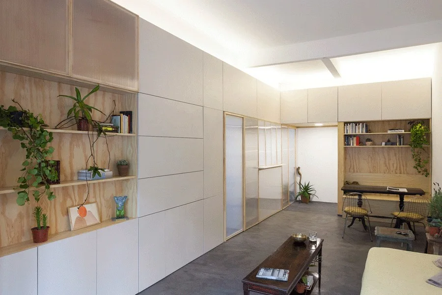 A modern living room features floor-to-ceiling custom storage cabinets with plywood shelving niches, a translucent sliding divider, and cove lighting.