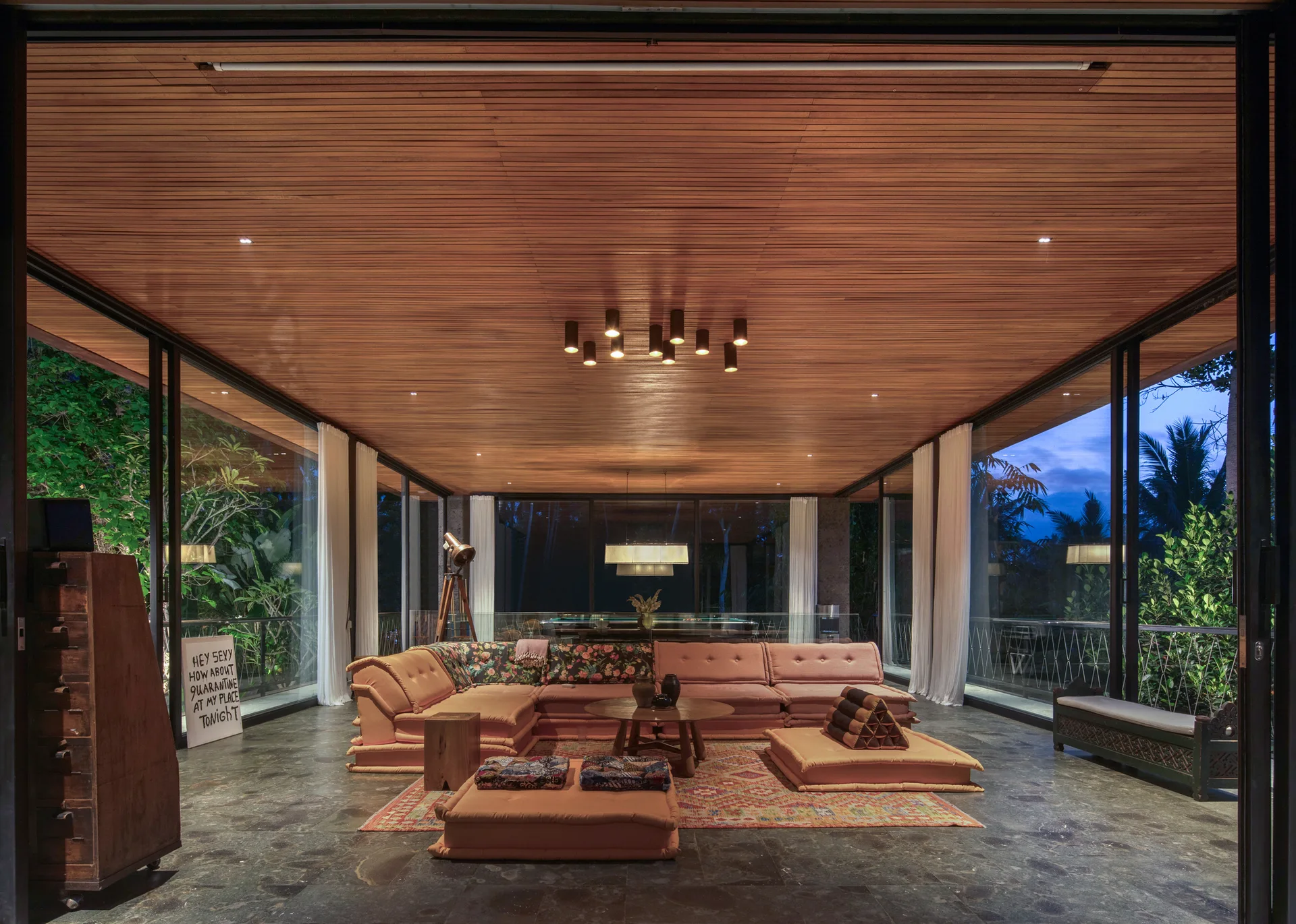 A spacious living room features a pink modular sectional, matching floor cushions, a wood slat ceiling, and glass walls opening to lush greenery.