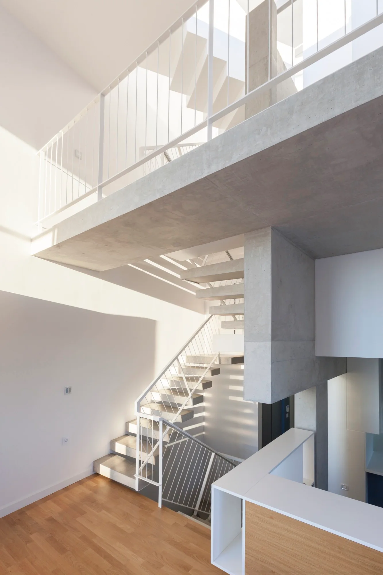 An interior view of a multi-level minimalist home featuring a floating concrete staircase with thin white metal railings and light wood floors.