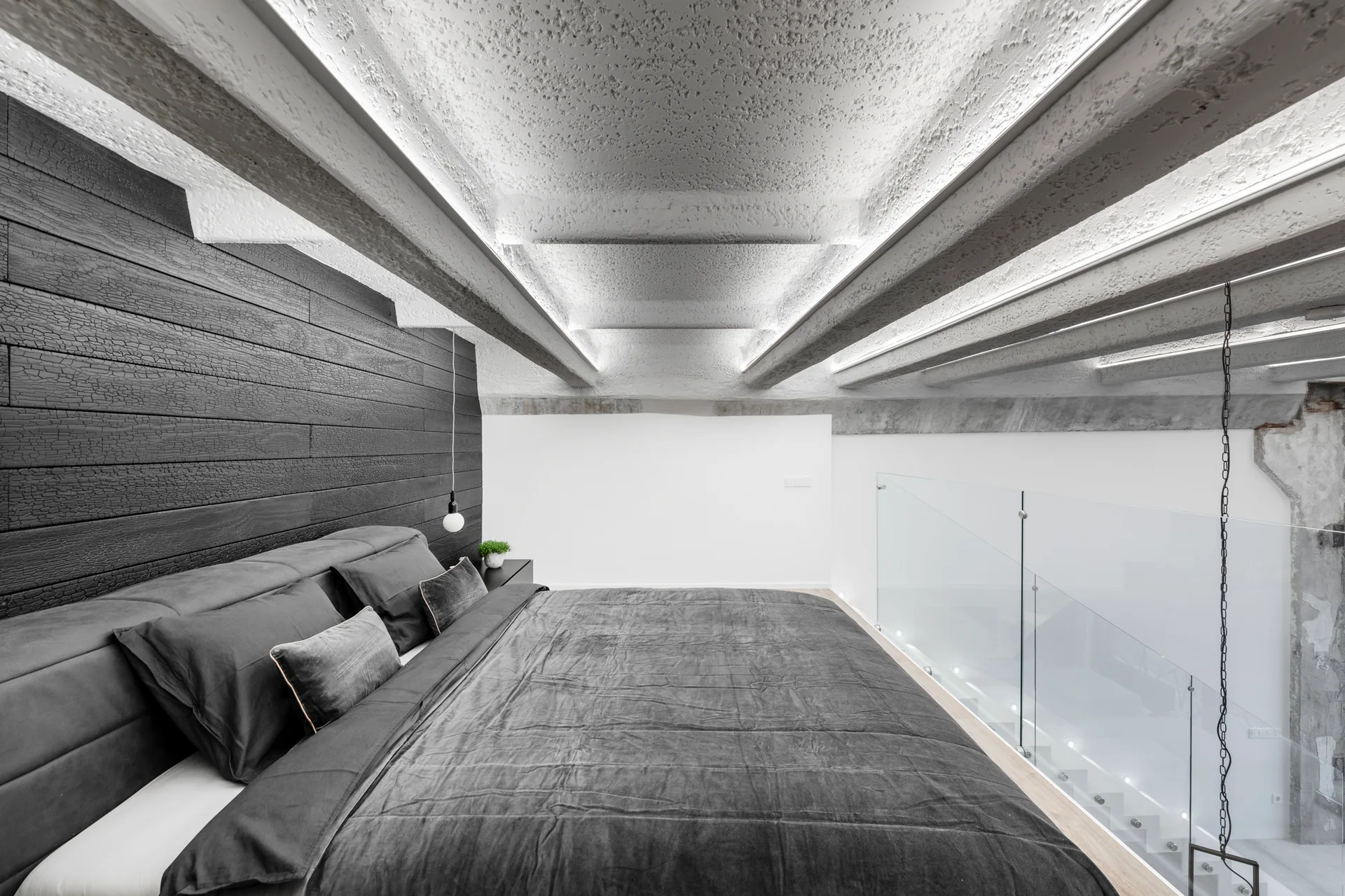 A minimalist loft bedroom features a dark grey bed set against a black textured wood wall beneath heavy white ceiling beams.