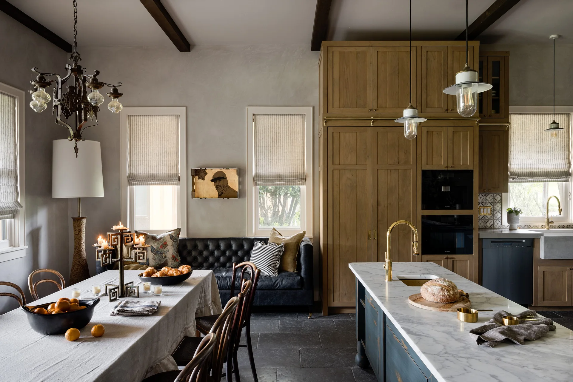 An open kitchen and dining space featuring a marble island, tall wood cabinets, a cloth-covered dining table, and a black leather tufted sofa.
