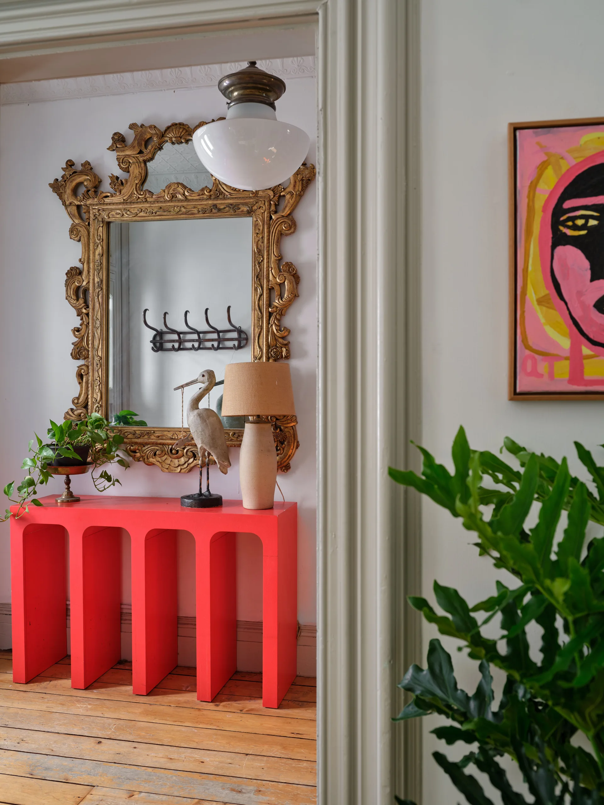 A bright coral entryway console table with a bird sculpture and lamp sits beneath an ornate gold mirror on natural wood floors.
