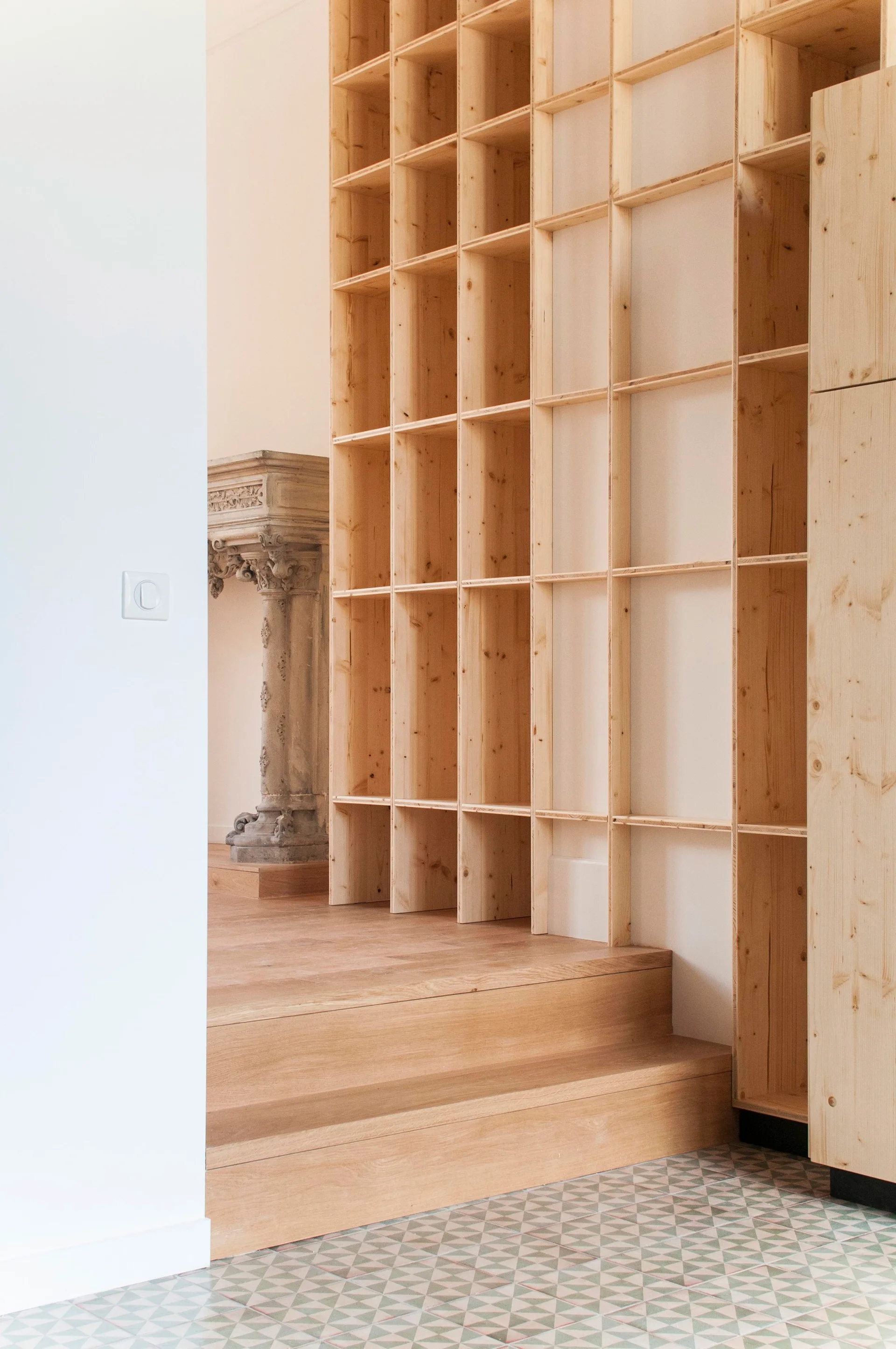 A tall light wood grid shelving unit stands next to short wooden stairs leading to an area with an antique carved stone column.