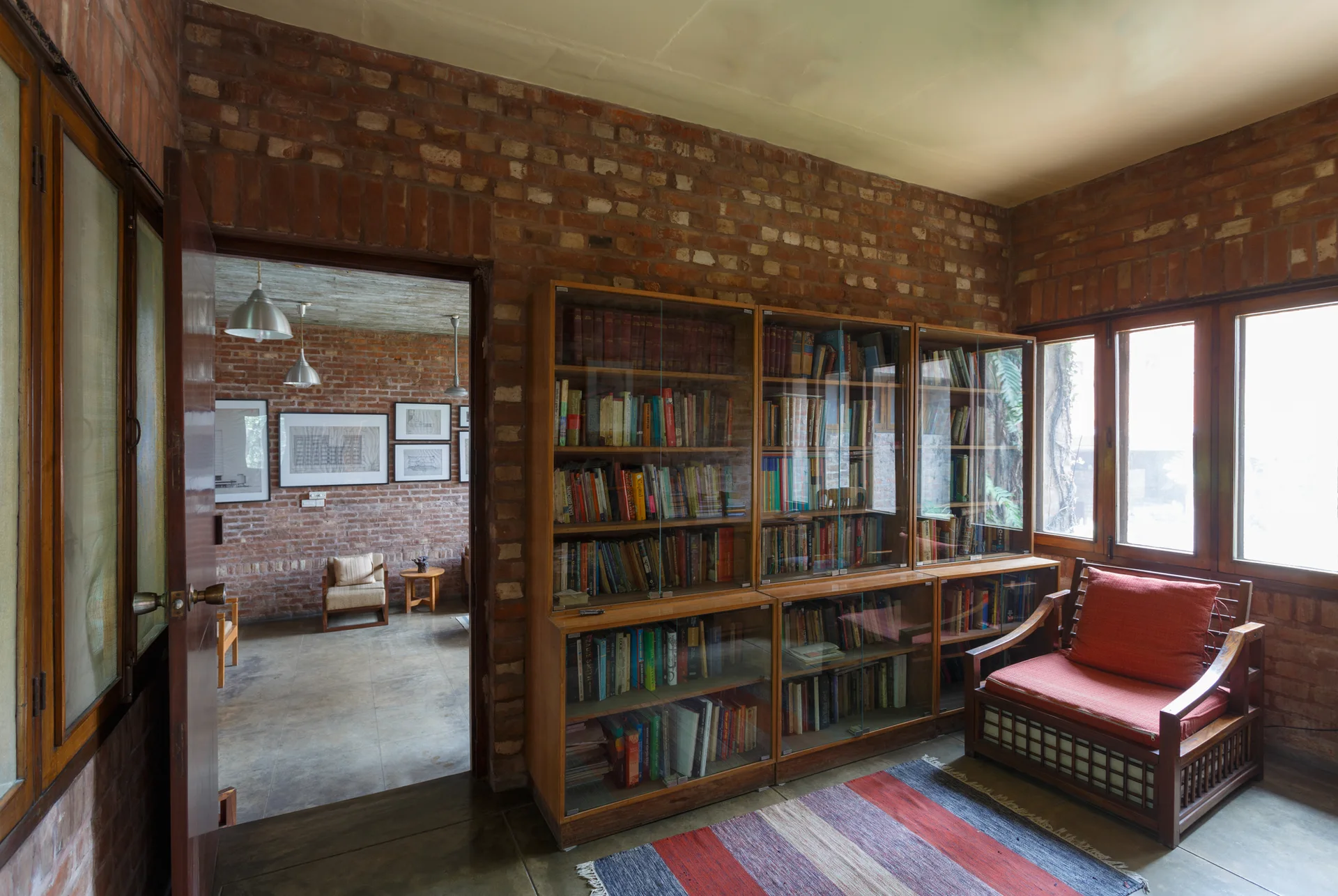 A home library featuring exposed red brick walls, a large glass-front wooden bookcase, and a wooden armchair with a red cushion.