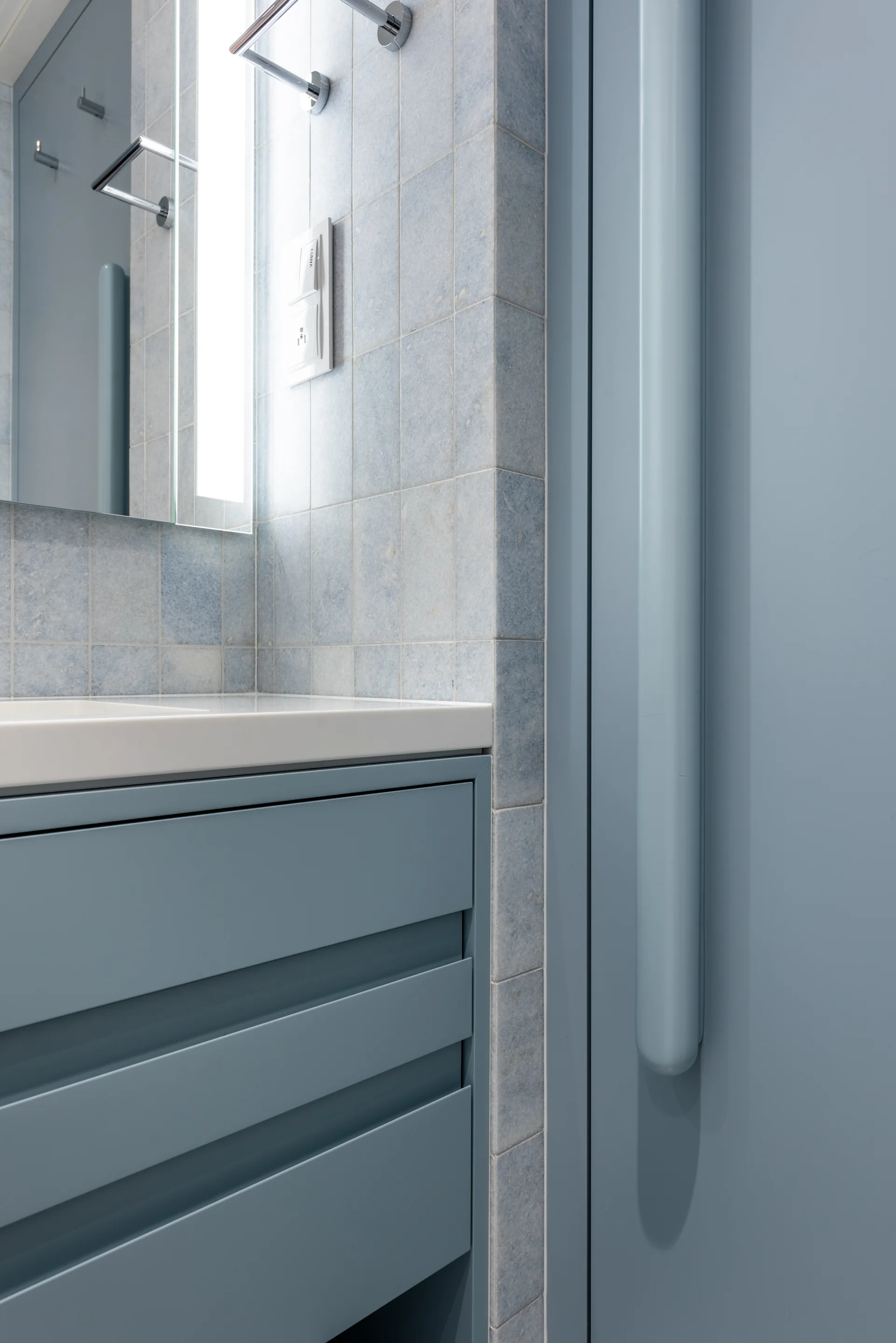 A close-up of a matte blue bathroom vanity with a white countertop set against a wall of light blue square ceramic tiles.