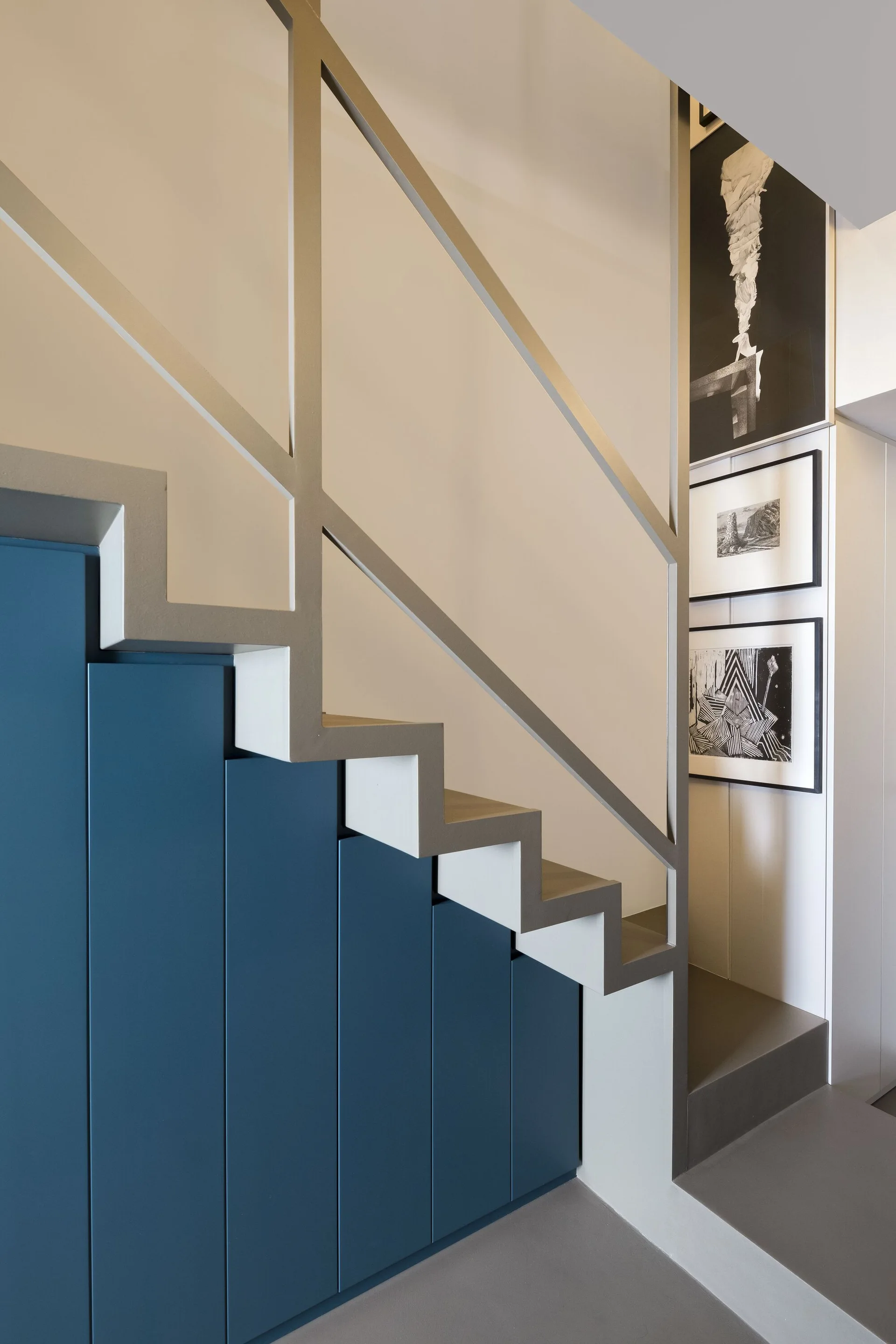 Modern grey zigzag staircase featuring custom blue cabinets fitted underneath, a geometric metal handrail, and black and white art on the adjacent wall.