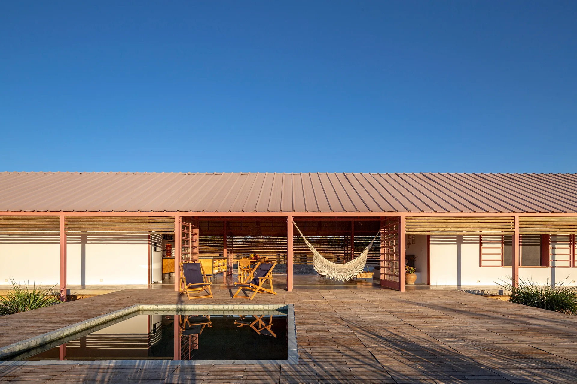 A wide wooden deck features a sunken square pool in front of an open-air house with a dusty pink steel roof frame and a hanging white hammock.