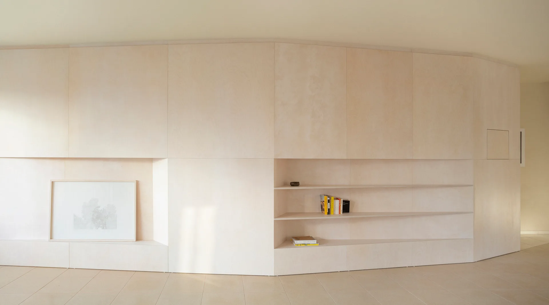 A minimalist wall clad in light plywood panels features integrated horizontal shelving and a recessed niche displaying framed art.