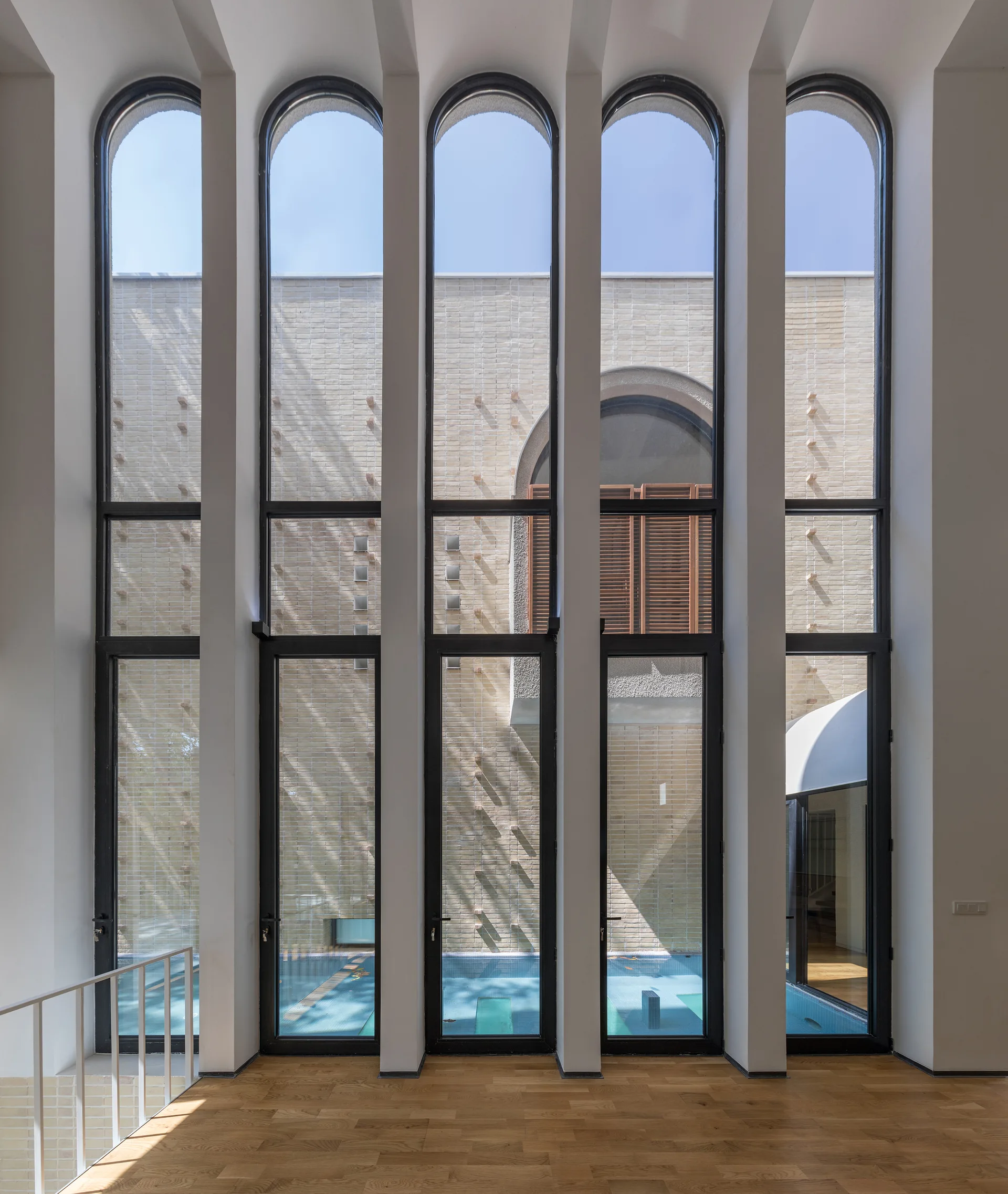 Tall arched floor-to-ceiling windows with black metal frames look out onto a courtyard swimming pool from a room with light hardwood floors.