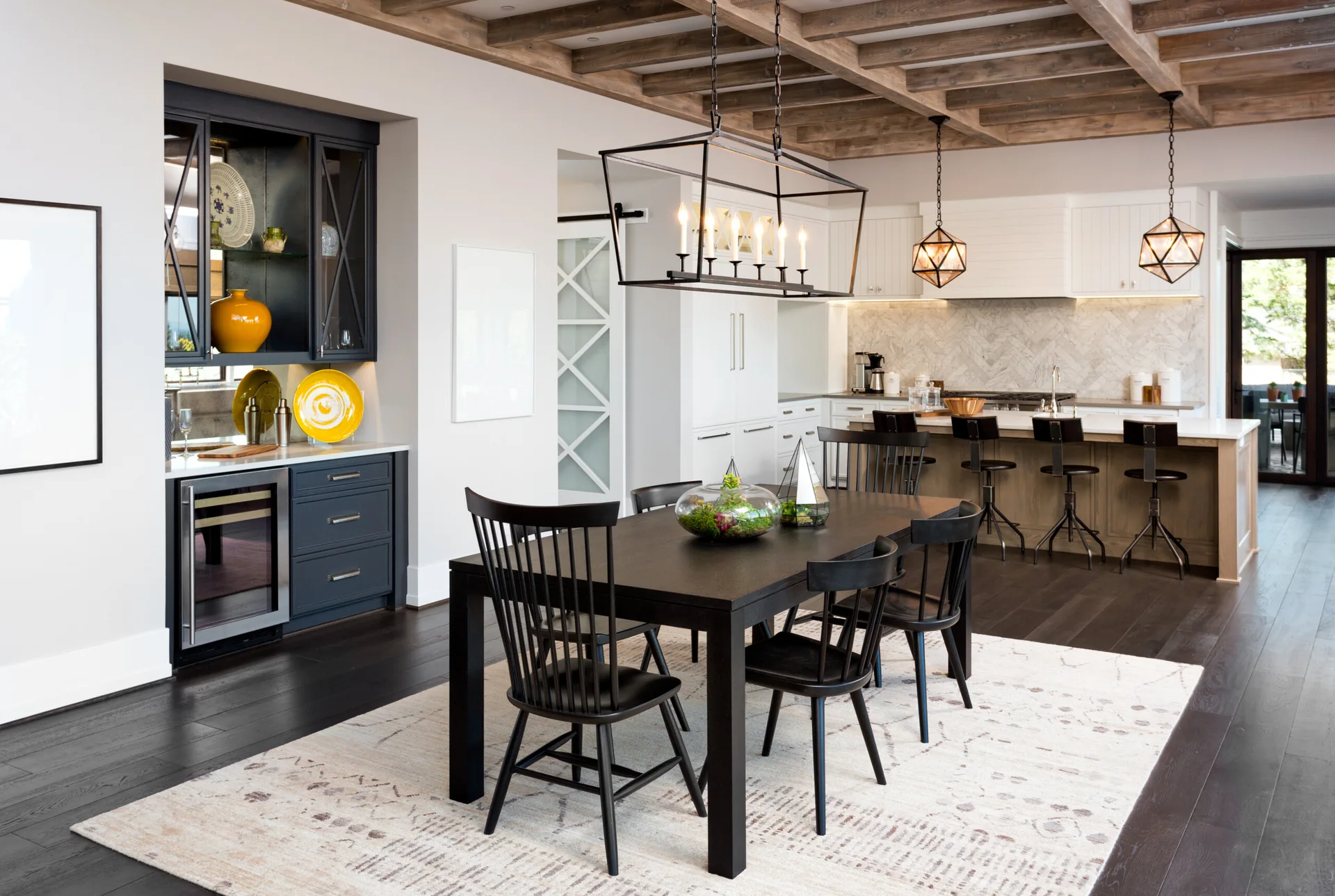 A transitional dining room with a black wooden table, spindle chairs, a light-colored rug, and rustic exposed wood beams on the ceiling.