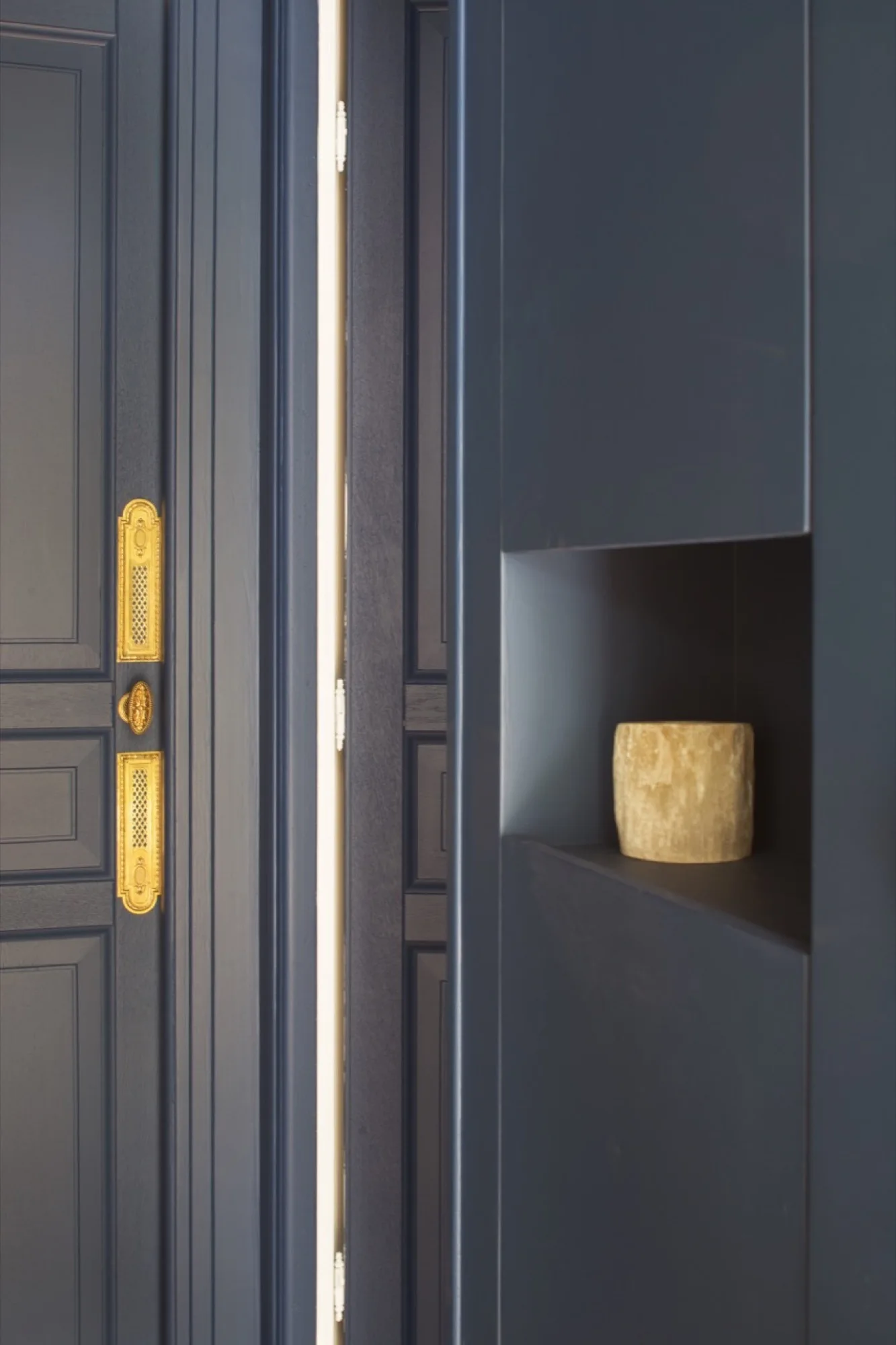 A dark blue paneled door with intricate gold brass hardware stands next to a matching blue wall with a small recessed square niche holding a vessel.
