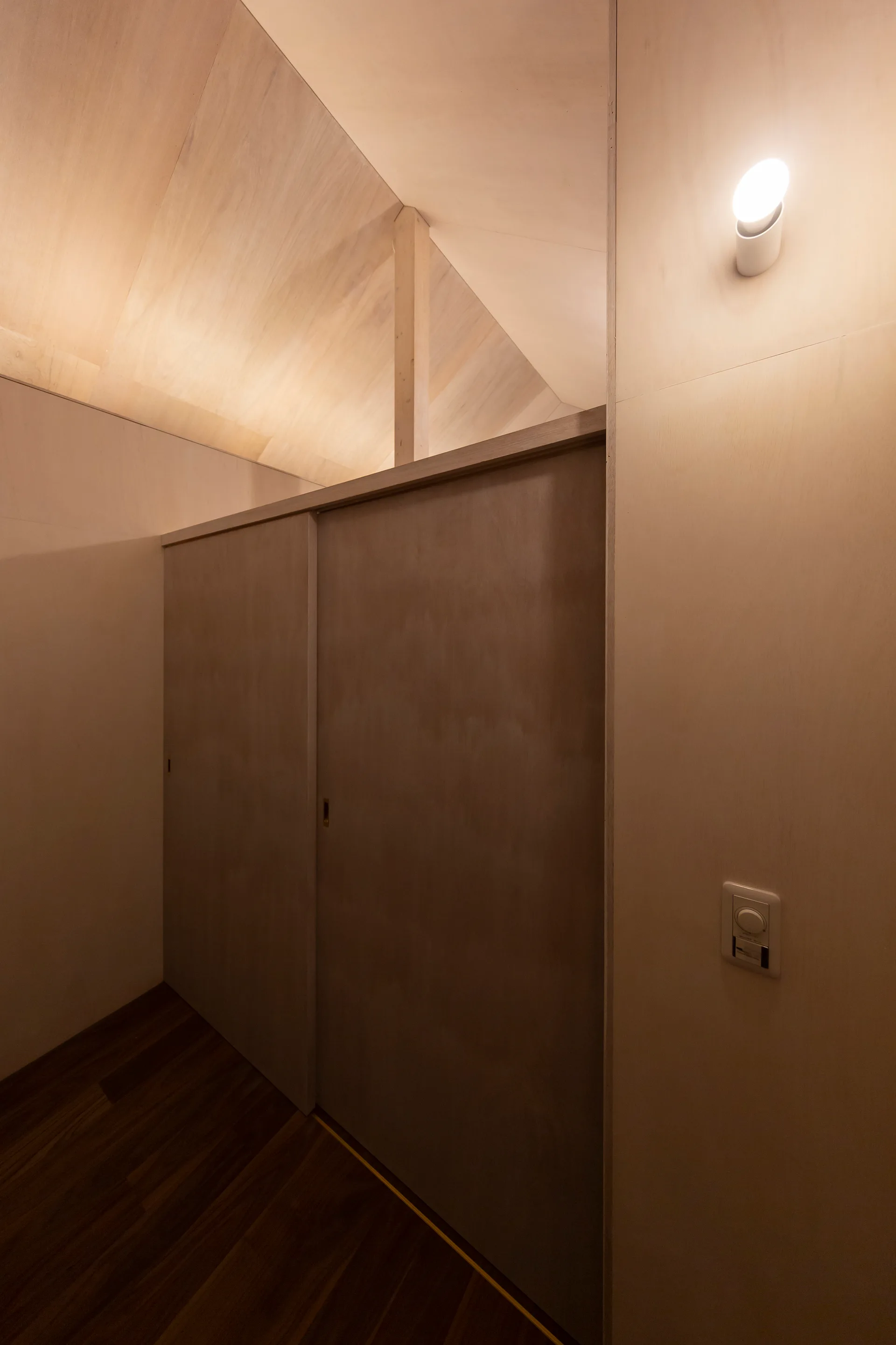 A minimalist hallway features light plywood walls and a vaulted ceiling, with recessed sliding doors and a single lit wall sconce.