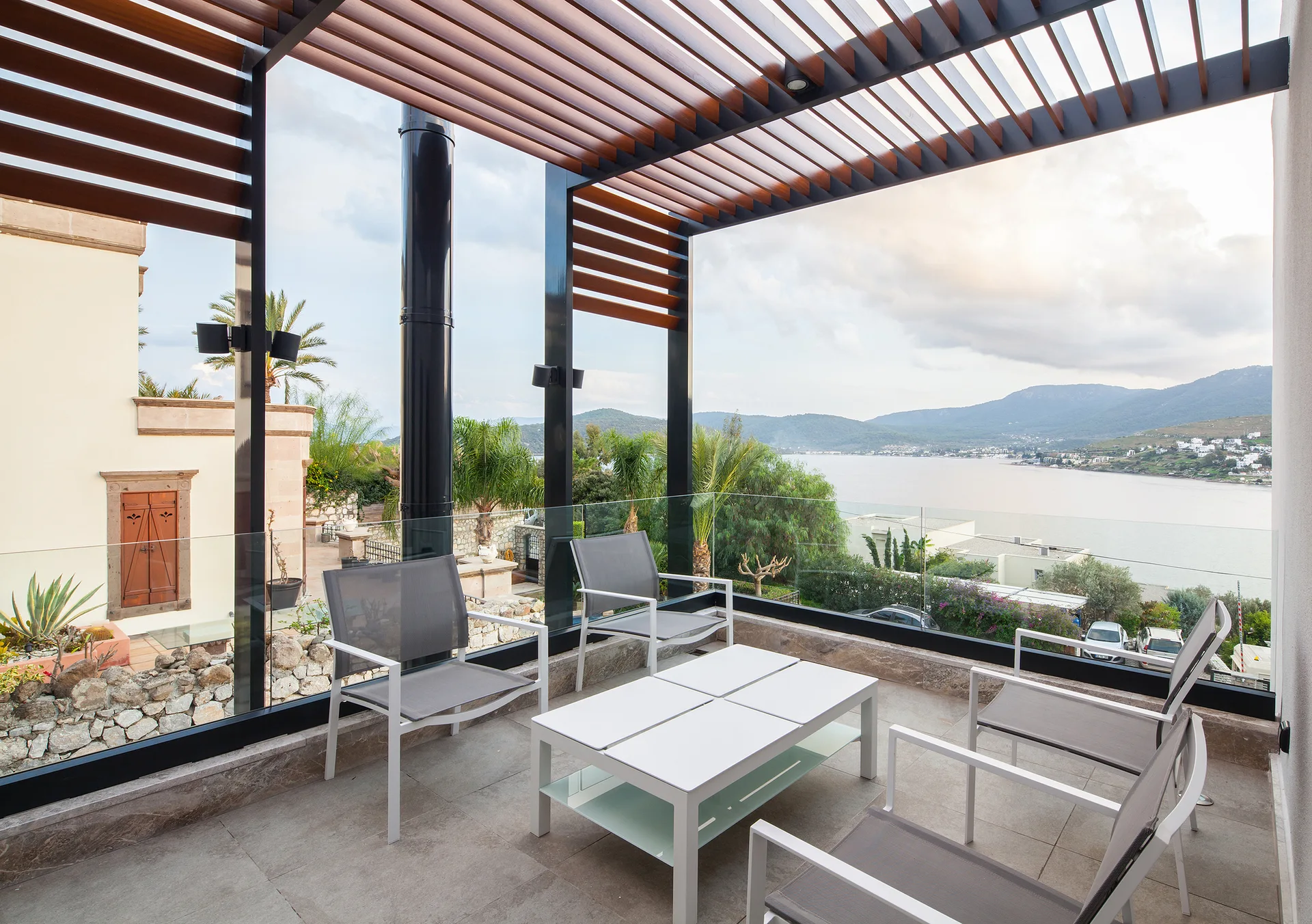 A modern outdoor terrace with a wood-slatted pergola roof, grey patio chairs around a white table, and a glass railing overlooking a bay.