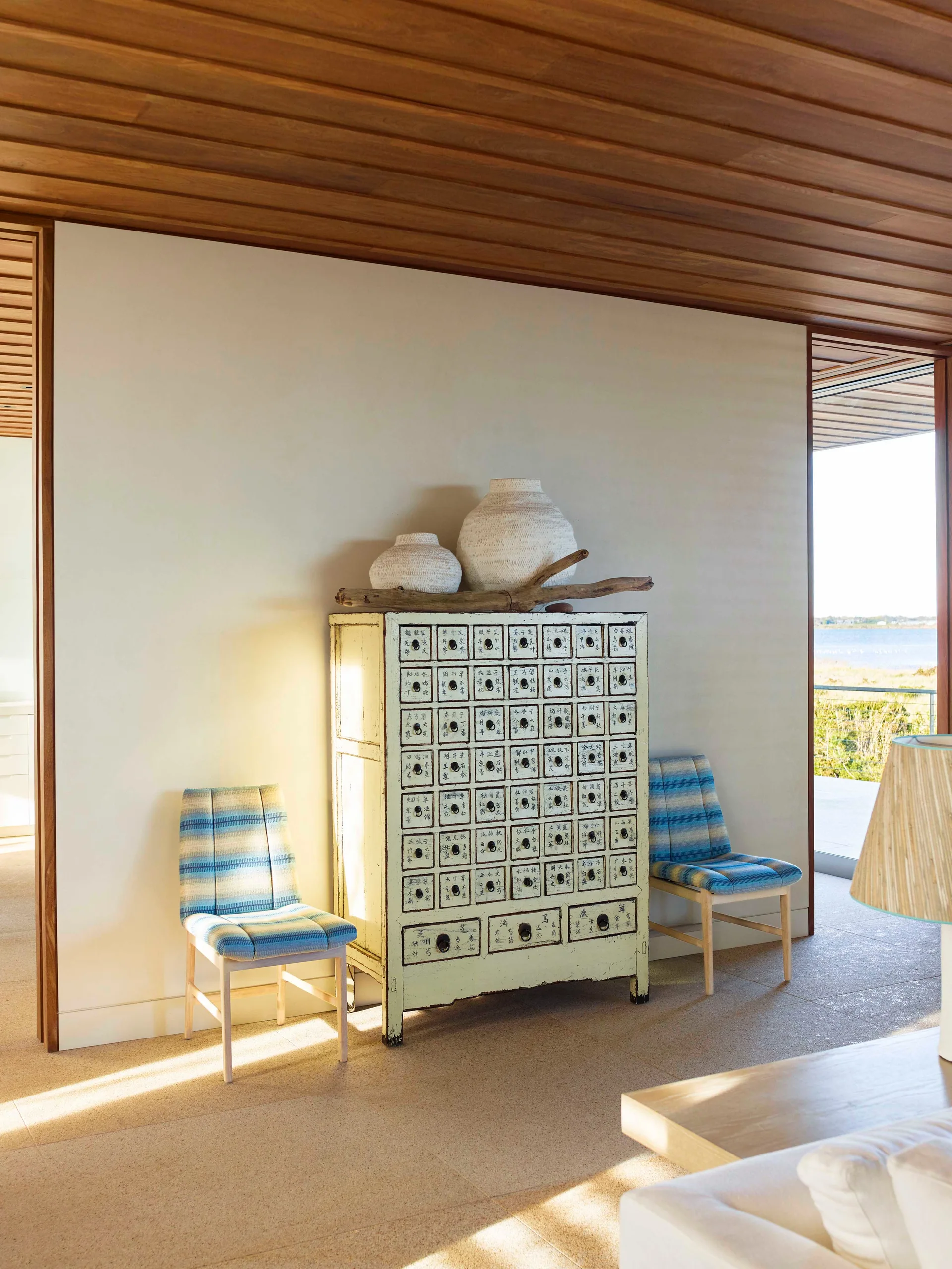 A cream apothecary chest with labeled drawers stands against a wall beneath a wood ceiling, flanked by two blue-striped chairs.