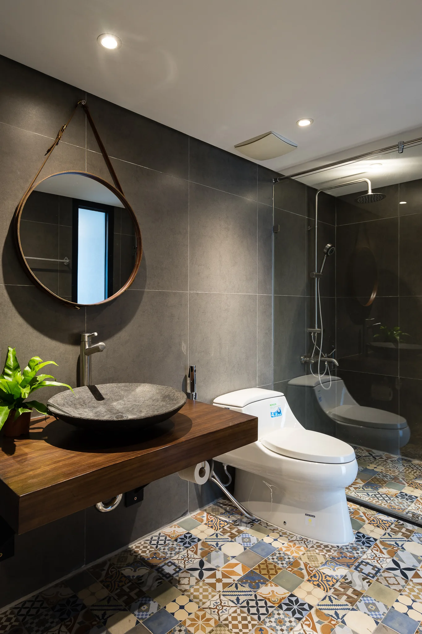 A contemporary bathroom features a floating wood vanity with a dark stone vessel sink, a round mirror on a strap, and colorful patterned floor tiles.