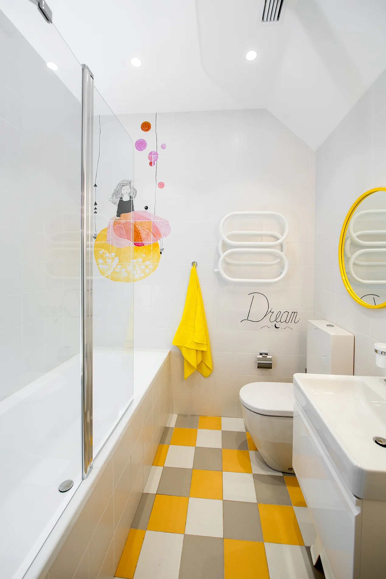 A bathroom features a yellow and grey checkered tile floor, a glass-enclosed bathtub, a white vanity with a yellow round mirror, and wall decals.