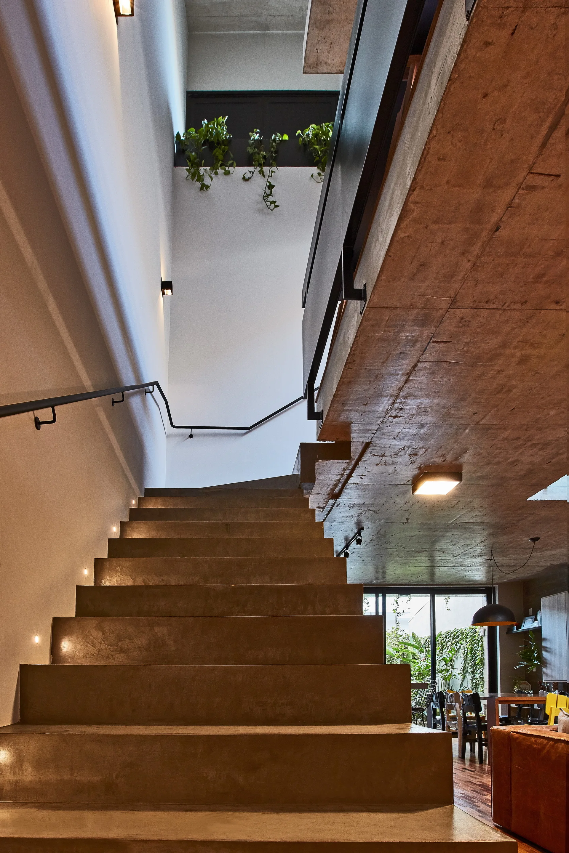 An industrial concrete staircase with a black metal handrail rises against a white wall illuminated by small step lights.